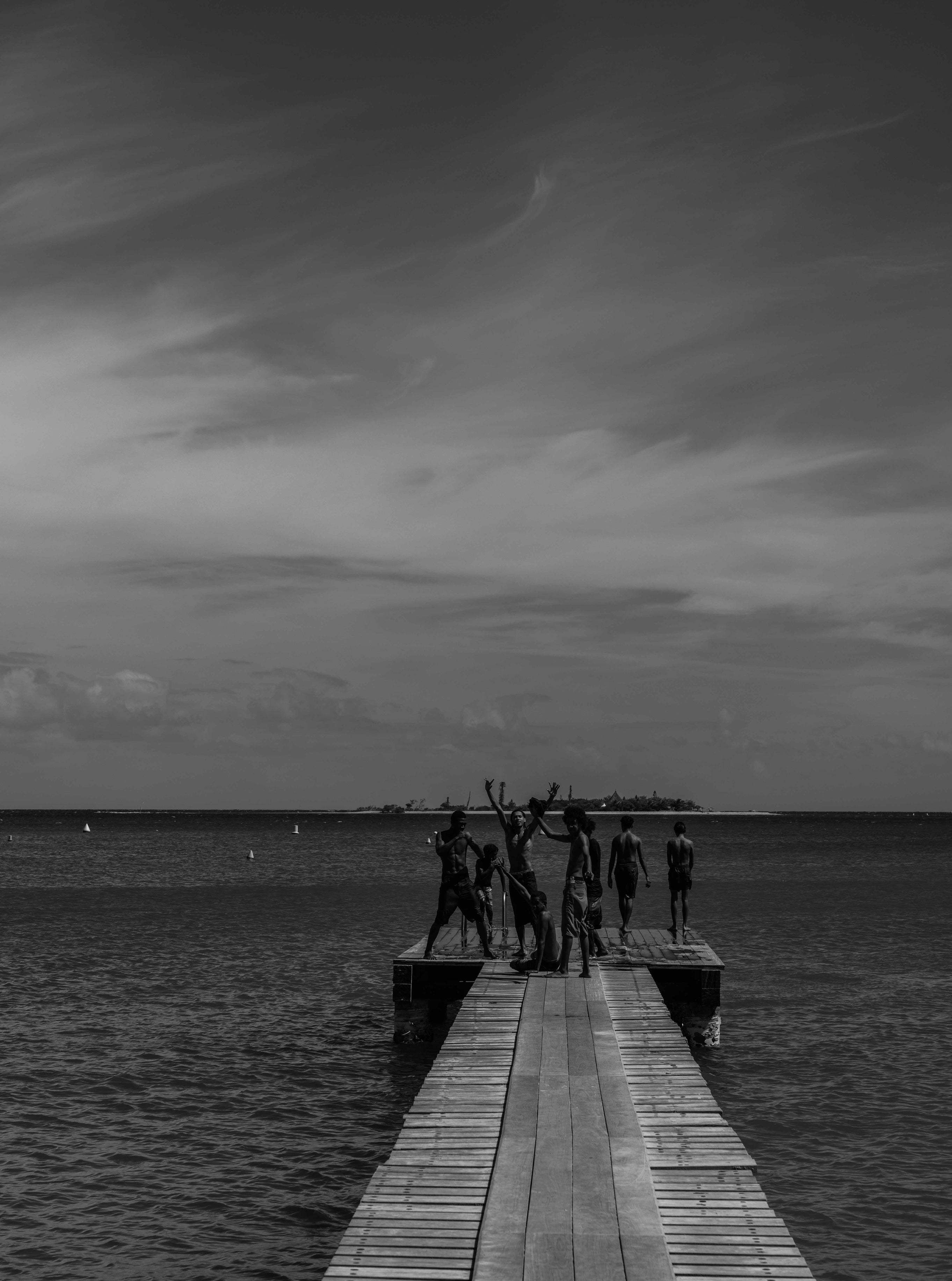 Black and white photograph of a group of kids standing at the end of a long wooden jetty extending into calm ocean water under a dramatic sky