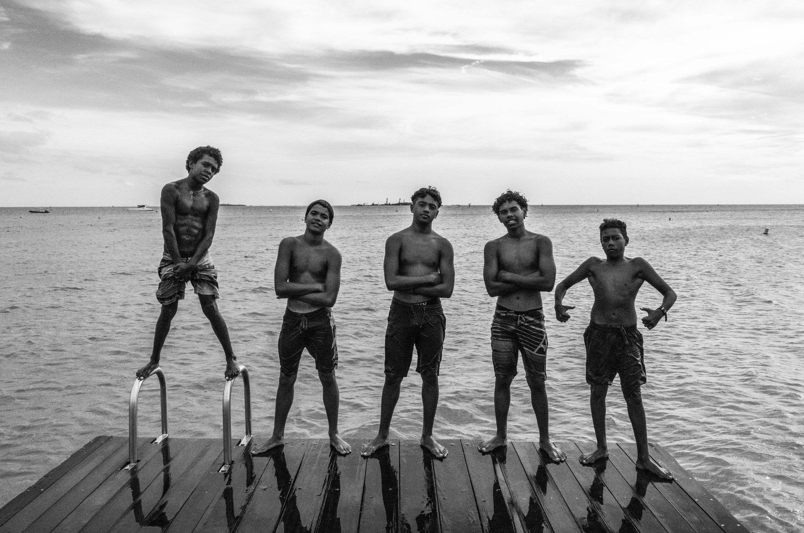 Black and white group portrait of five young men standing in a row on a wooden dock with ocean and sky behind them