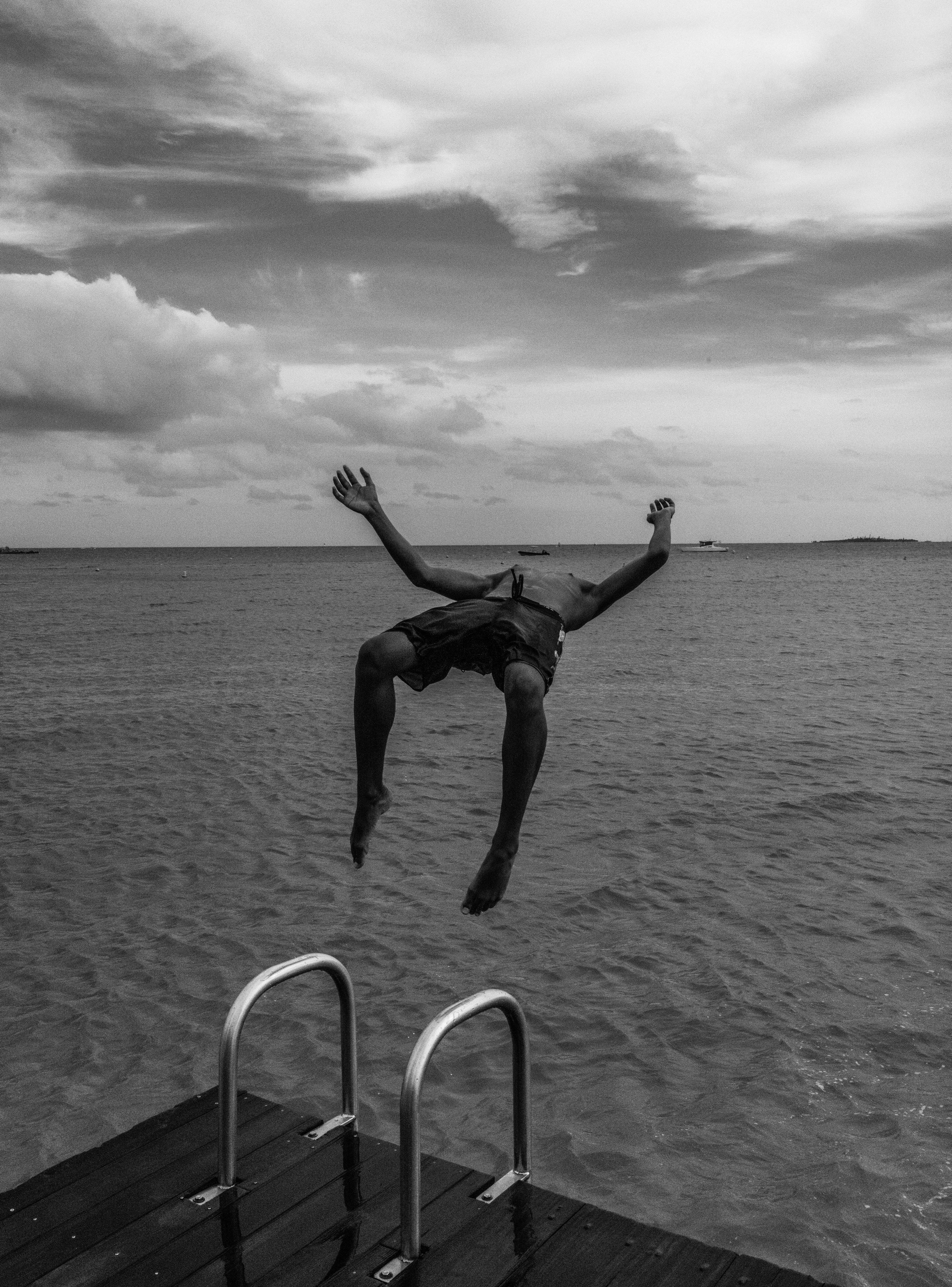 Black and white photograph of a boy mid-backflip off a wooden jetty with a metal ladder visible and ocean stretching to the horizon