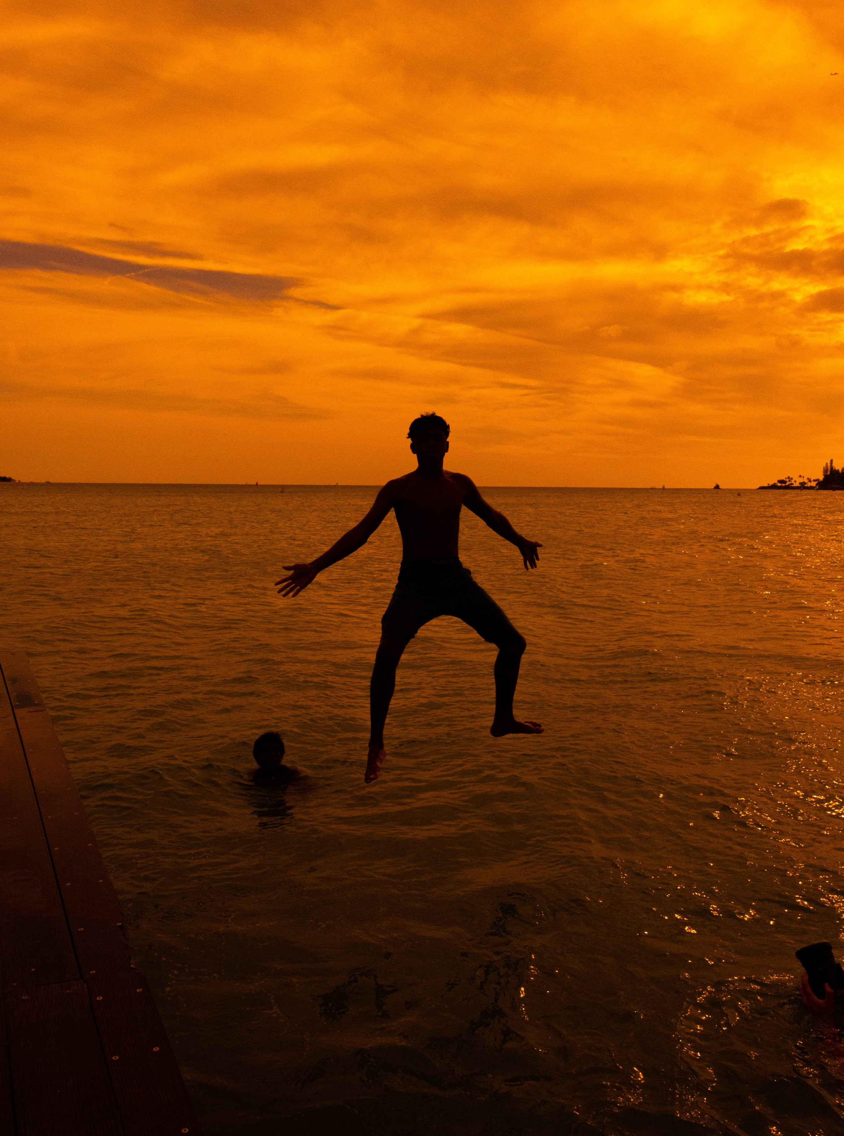 Silhouetted boy mid-jump into the ocean against a vivid orange sunset sky with another boy swimming in the golden water below