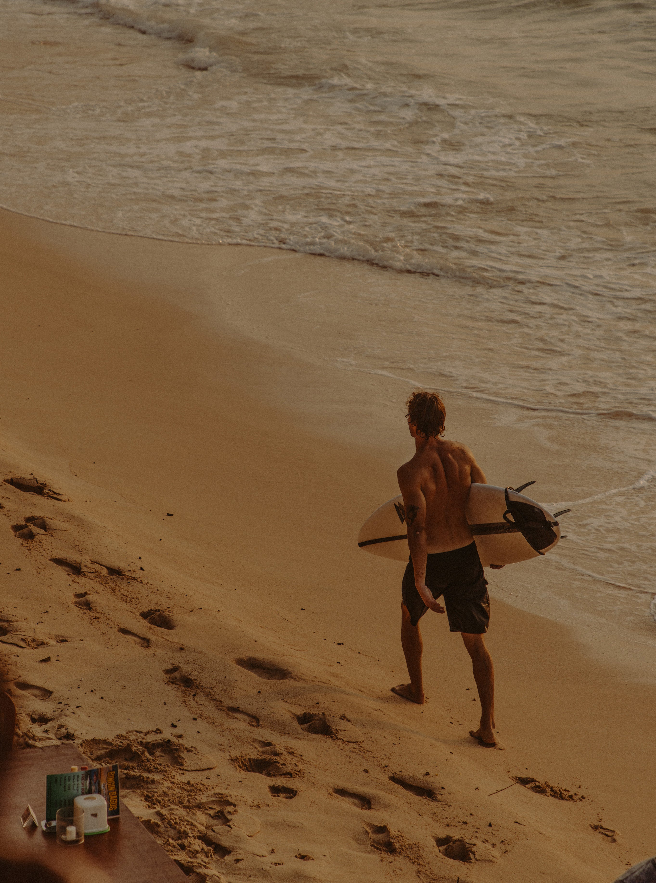 Rear view of a shirtless surfer carrying a surfboard walking toward the waves on golden sand at dusk