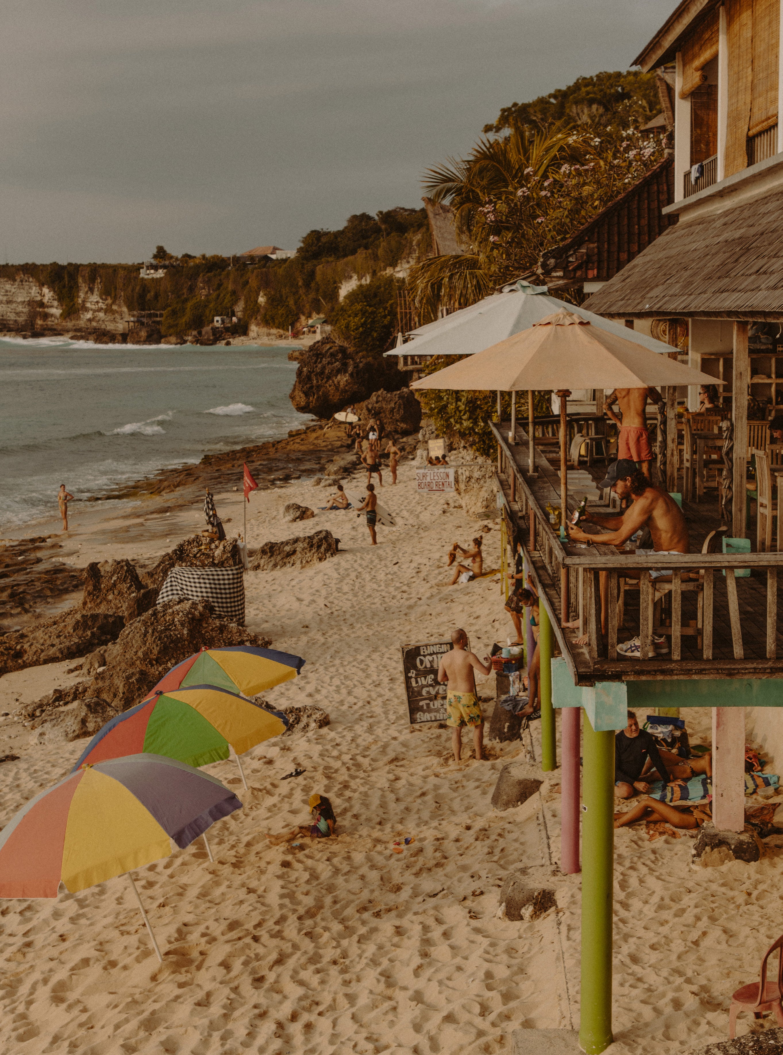 Elevated view of a busy beach club with colourful umbrellas, beachgoers and a wooden bar structure set against rocky cliffs and ocean