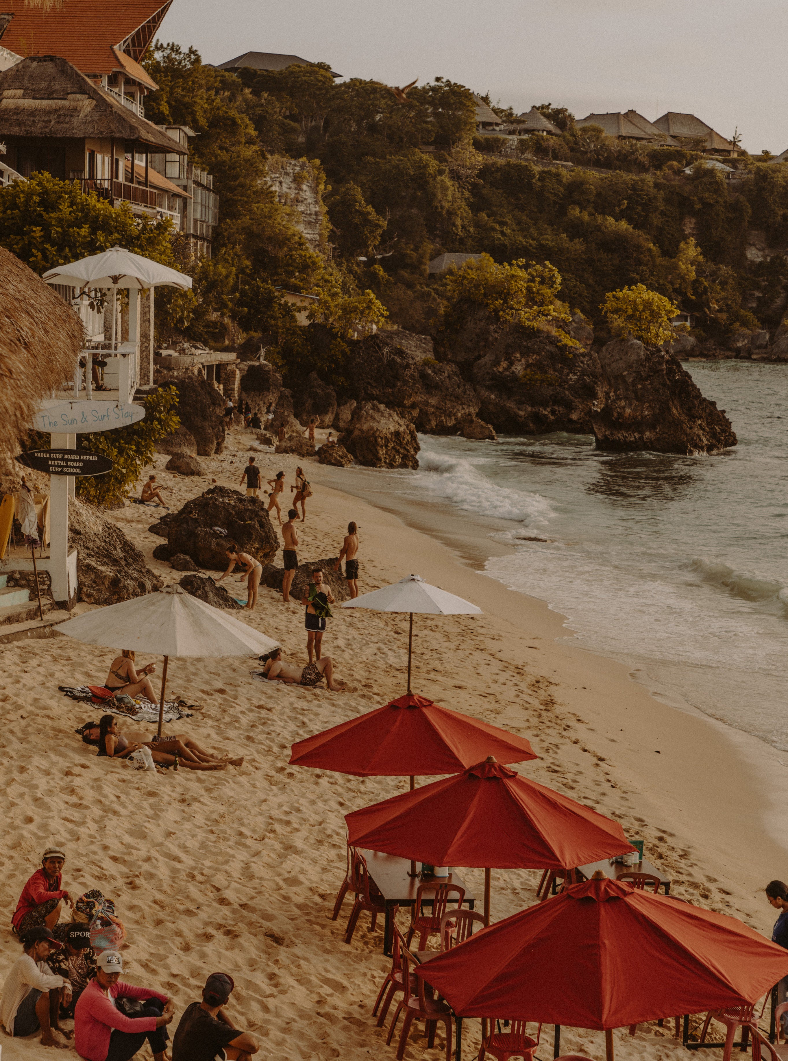 Elevated view of a sandy beach cove with red umbrellas, beachgoers and a clifftop bar surrounded by tropical vegetation and rocky headlands