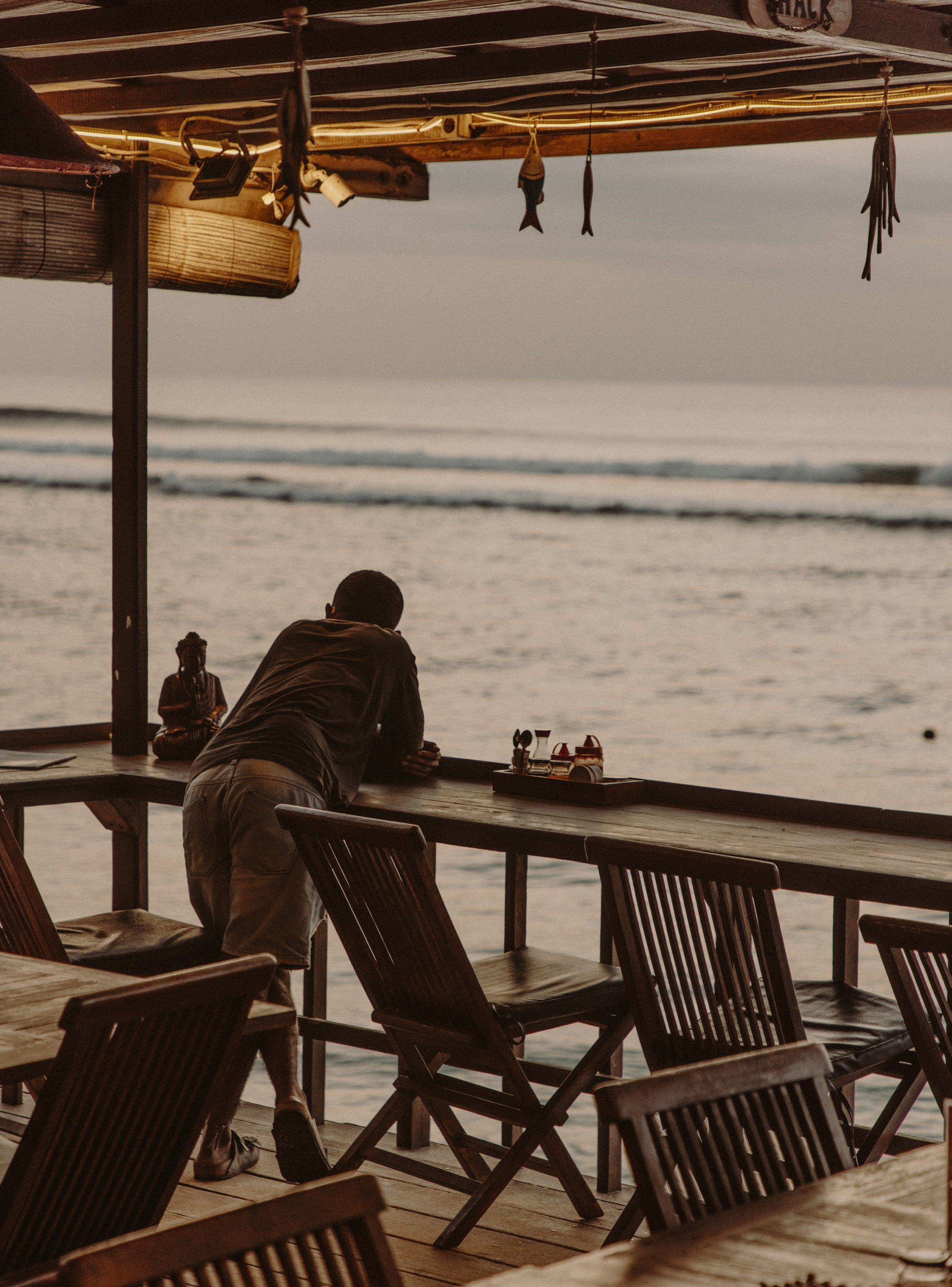 Silhouetted figure sitting alone at a wooden beachside bar looking out at the ocean at golden hour with small wooden fish hanging from the roof