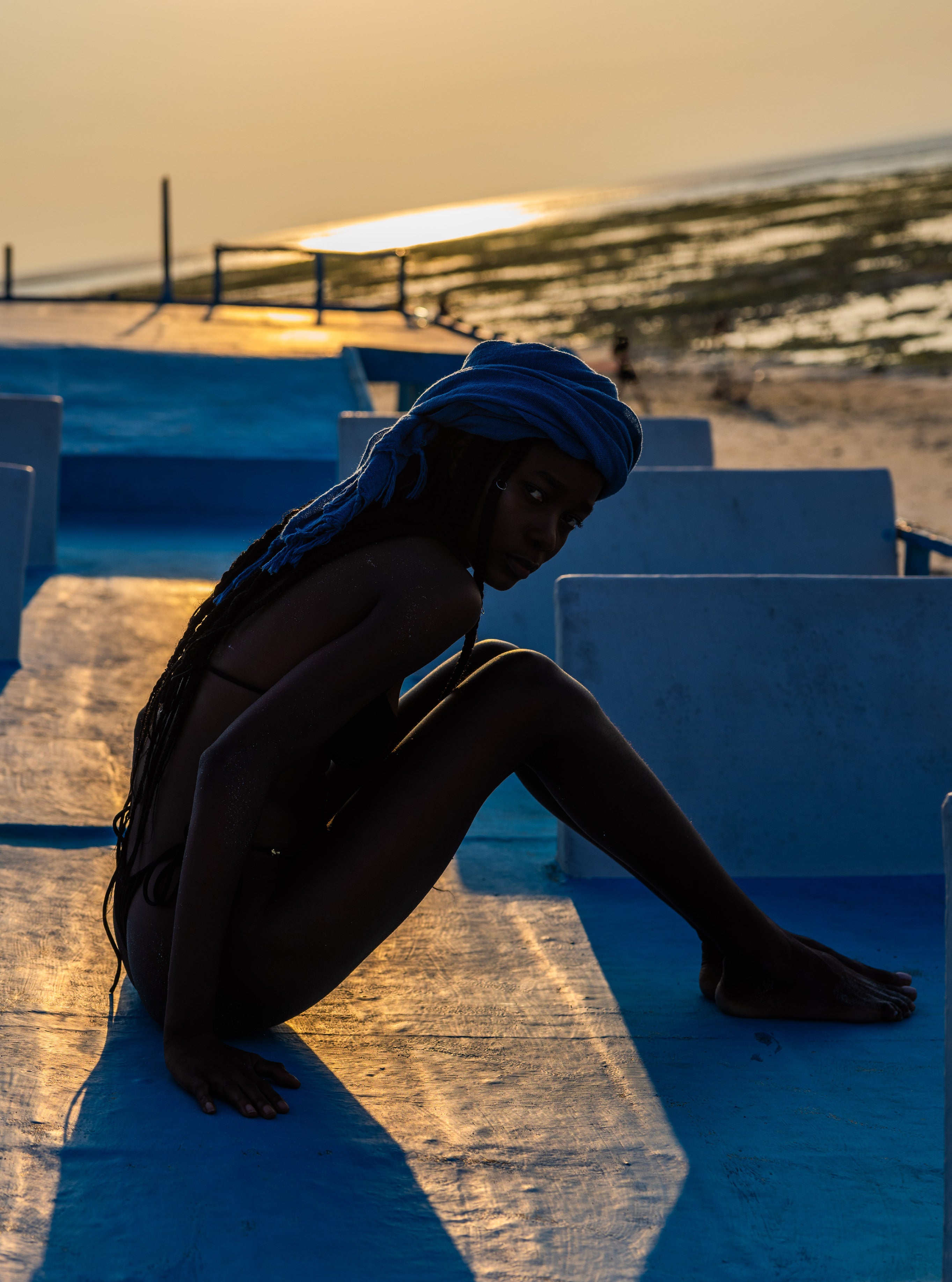 Woman wearing a blue headwrap sitting on blue-painted concrete steps at the beach at dusk with golden sunset light on the horizon behind her