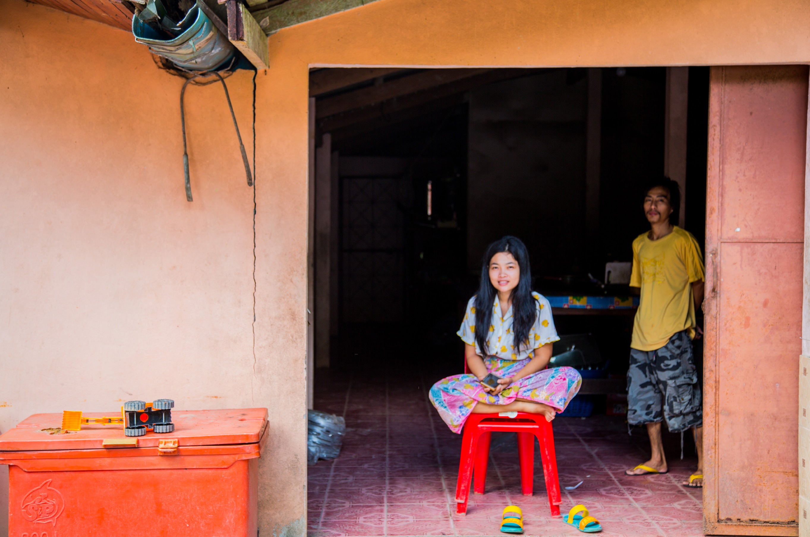 Young girl sitting on a small red stool in the wide doorway of a peach-walled village house with a man standing in the doorway behind her