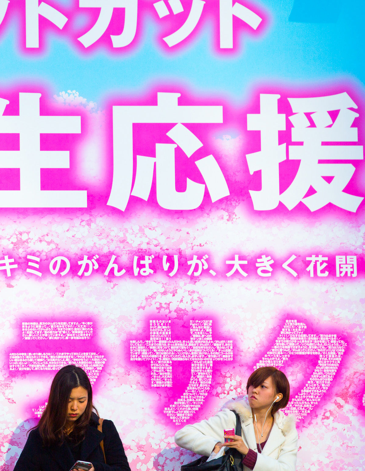 Two women stand beneath a bright pink billboard featuring cherry blossom graphics and Japanese text on a Tokyo street