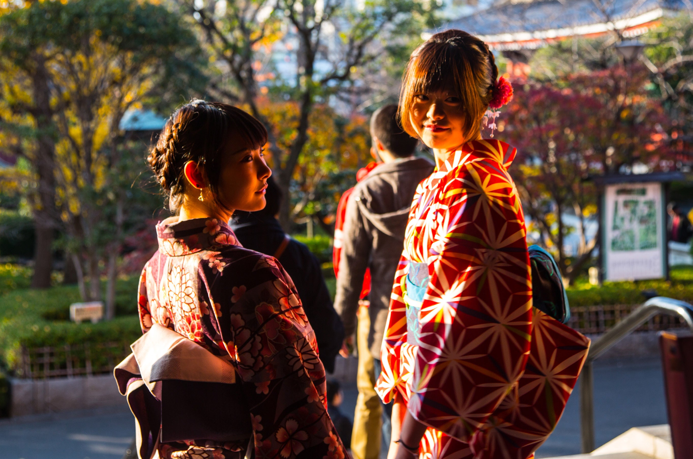 Two young women wearing colourful traditional kimonos, one dark floral and one red geometric, stand together in golden autumn light at a Tokyo temple garden
