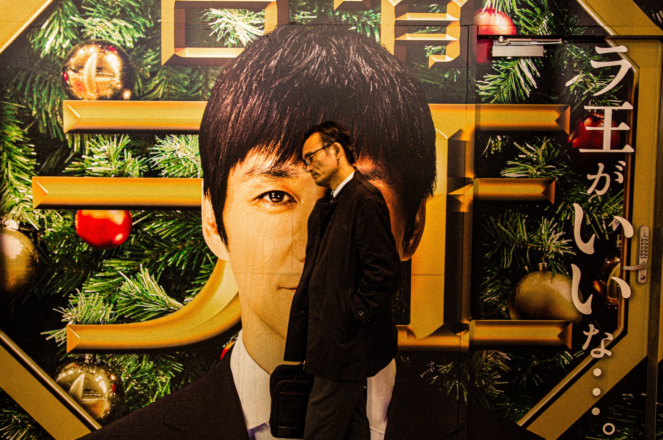 A man in a dark suit walks past a large illuminated Japanese billboard, his figure overlapping with the giant face on the advertisement in Tokyo at night