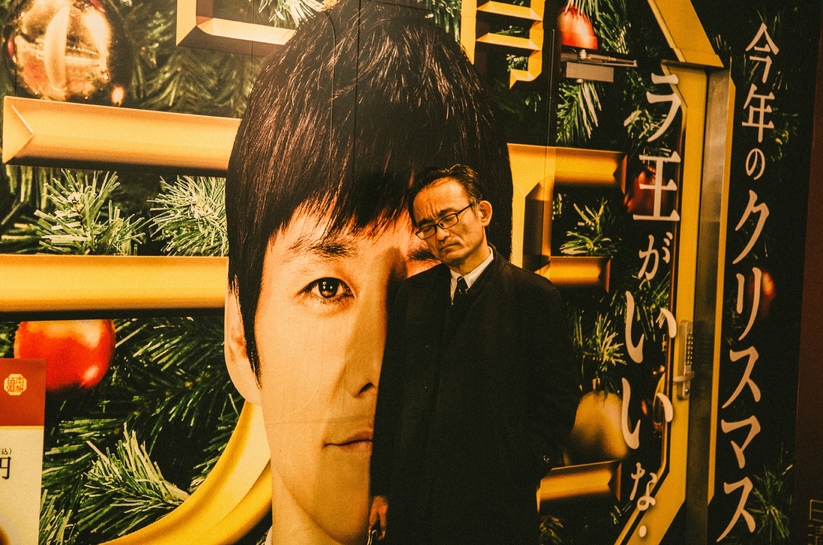 Tight crop of a man standing in front of a large illuminated Japanese billboard, his body overlapping the oversized face in the advertisement