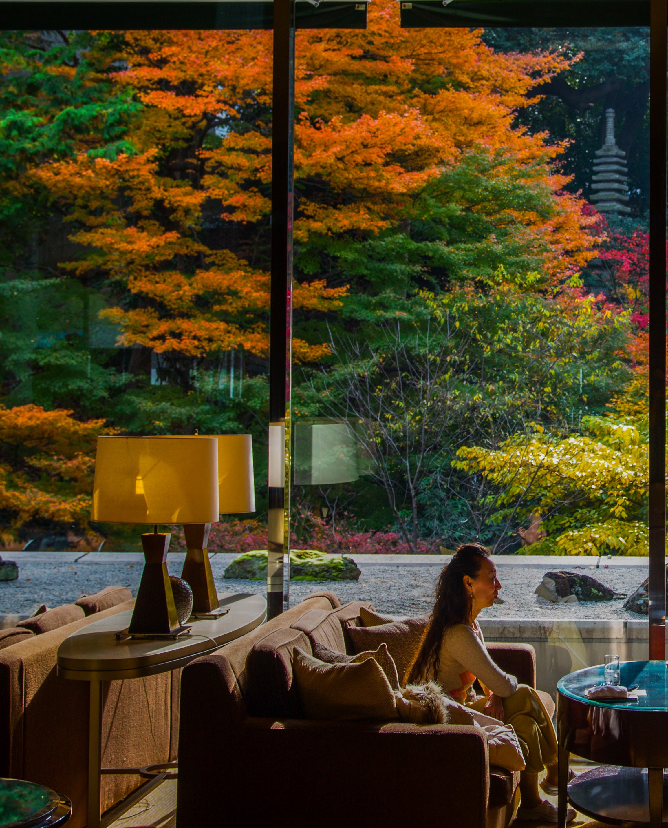 A woman sits in a lounge chair looking out through a large window at a Japanese garden ablaze with orange and red autumn maple trees in Kyoto