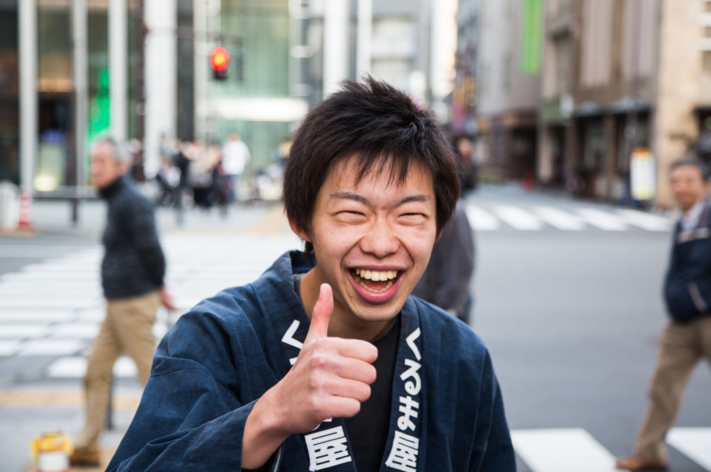 A young smiling rickshaw runner wearing a traditional dark blue happi coat gives an enthusiastic thumbs up on an Asakusa street in Tokyo