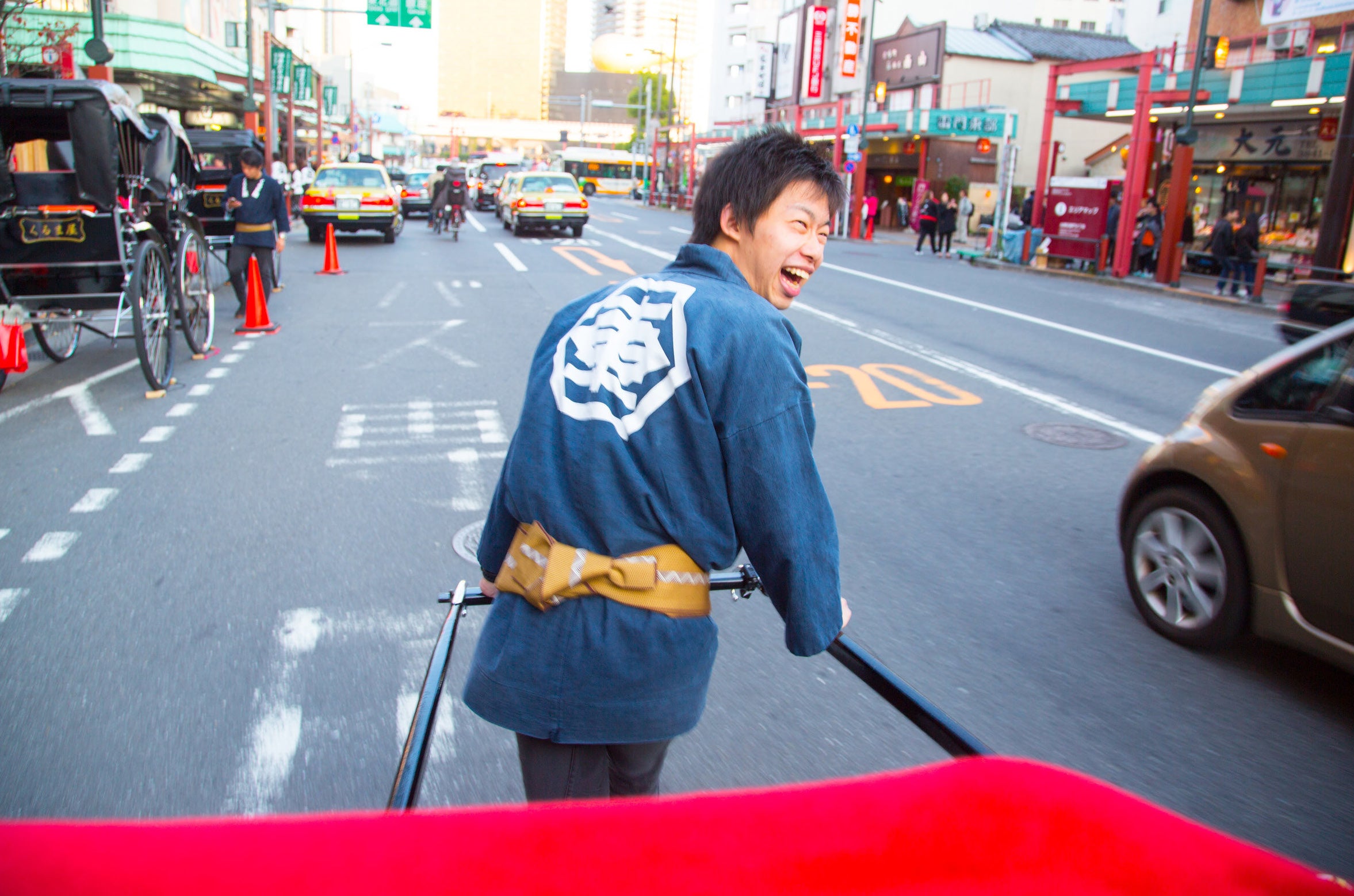 A laughing rickshaw runner in a traditional happi coat pulls passengers through the busy streets of Asakusa, Tokyo, shot from the passenger perspective with motion blur