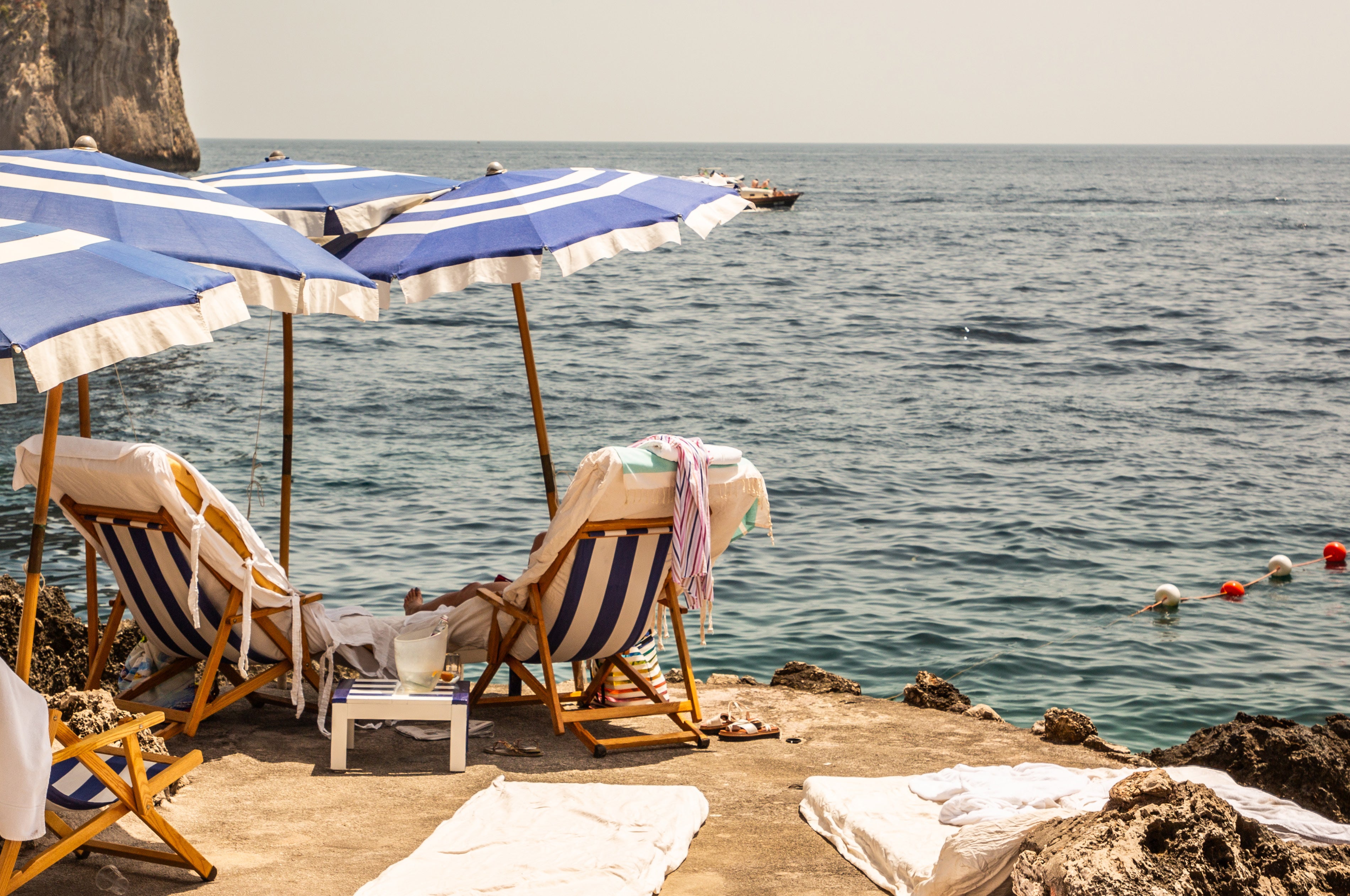 Two blue and white striped beach umbrellas shade wooden deck chairs on a rocky limestone platform overlooking the calm Mediterranean Sea in Capri, Italy