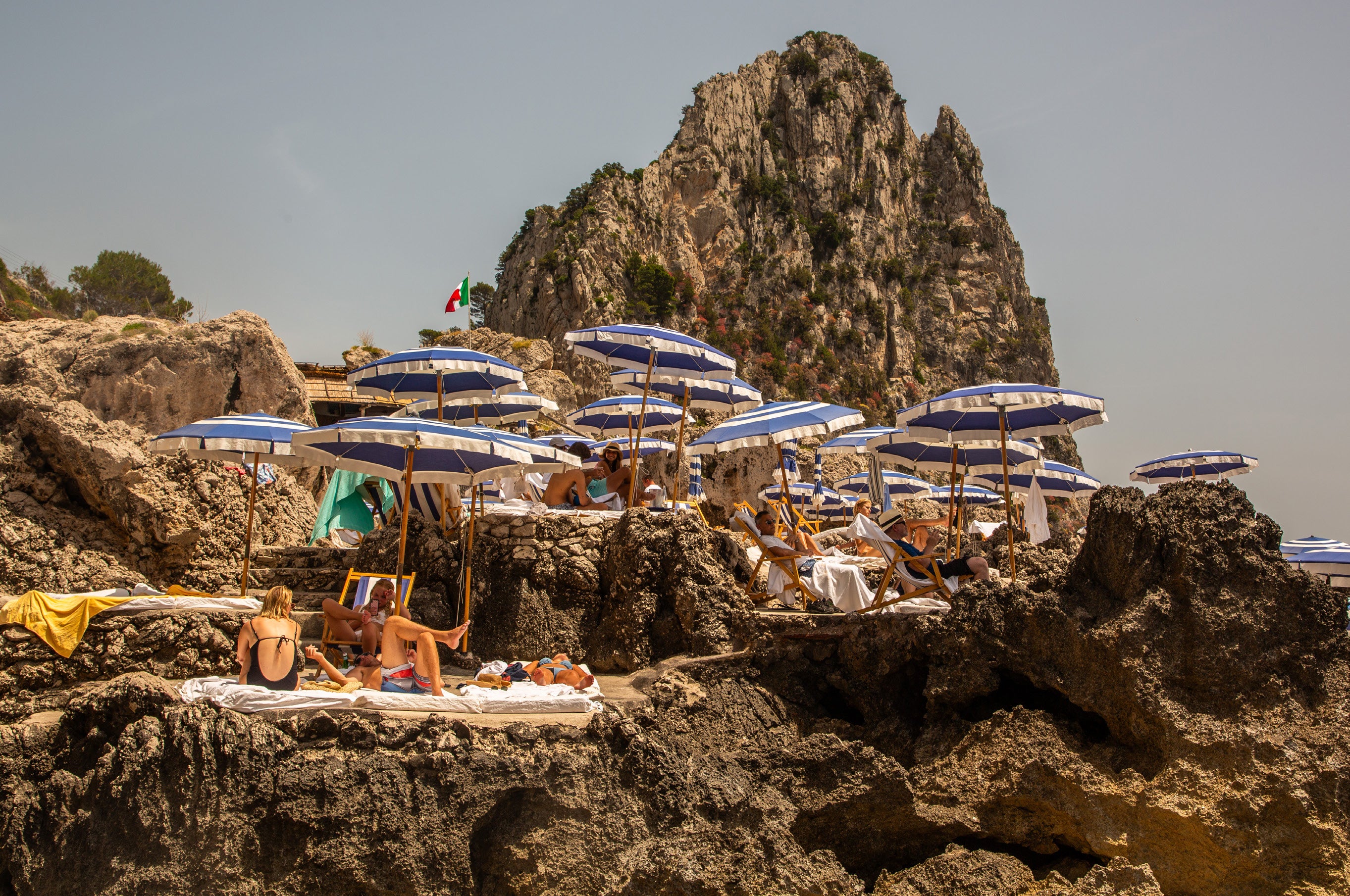 Sunbathers lounge on towels and deck chairs among blue and white striped umbrellas nestled into dramatic rocky cliffs at a beach club on Capri, Italy
