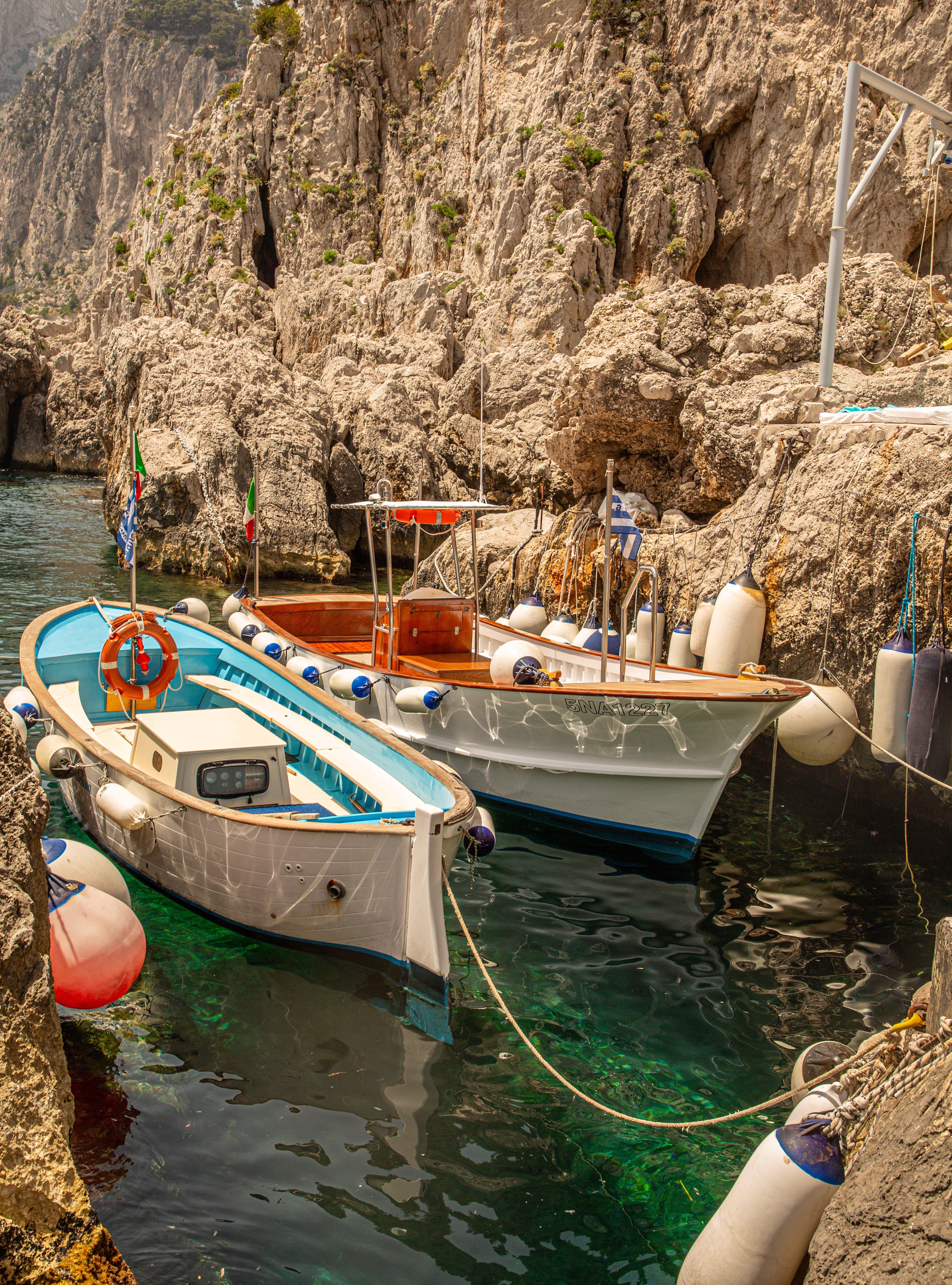 Several colourful traditional wooden boats moored together in crystal-clear emerald water at the base of tall rugged limestone cliffs on Capri