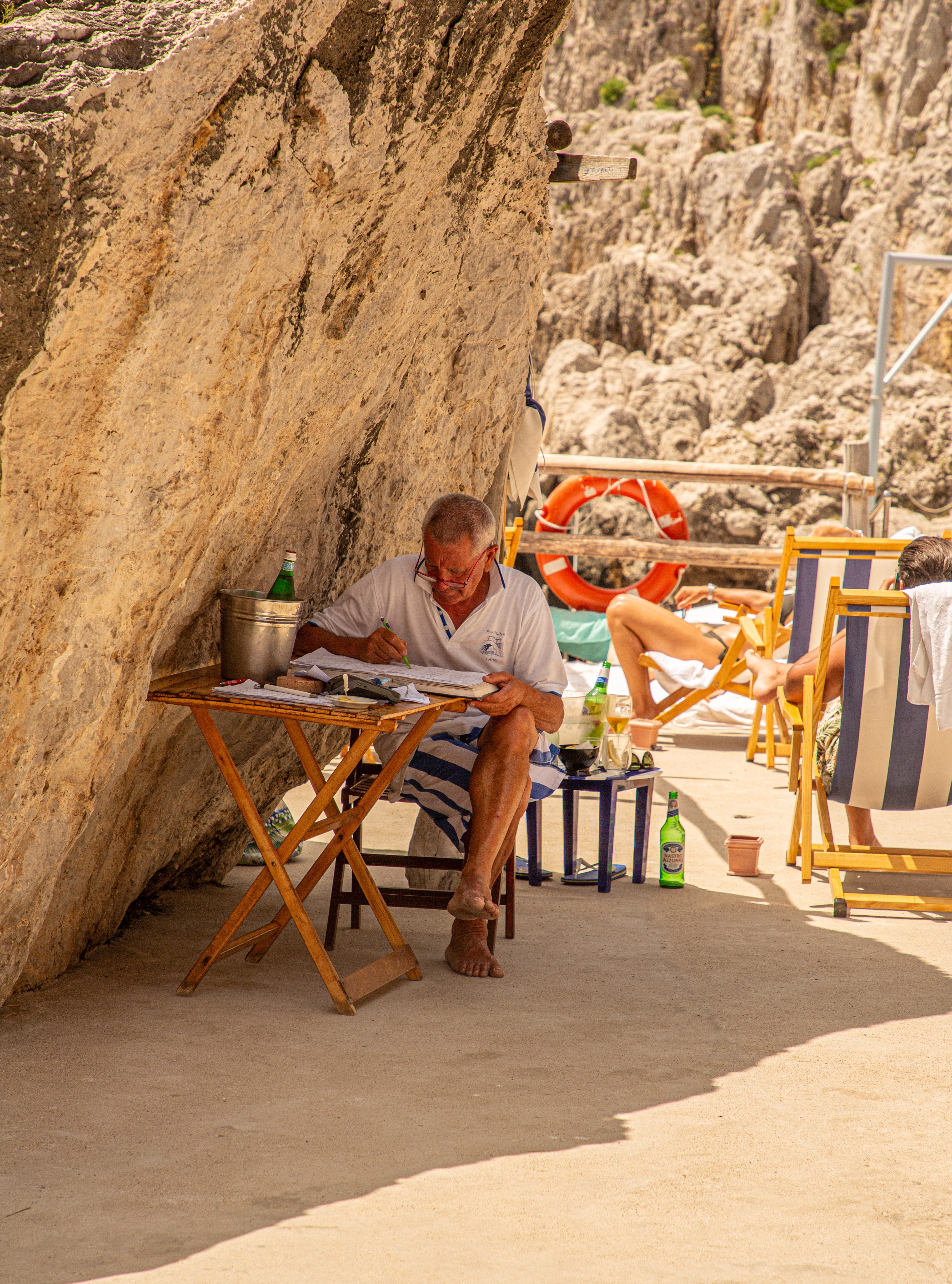 An older man in a white polo shirt sits at a small folding wooden table writing in a ledger beneath a sunlit limestone rock overhang at a Capri beach club