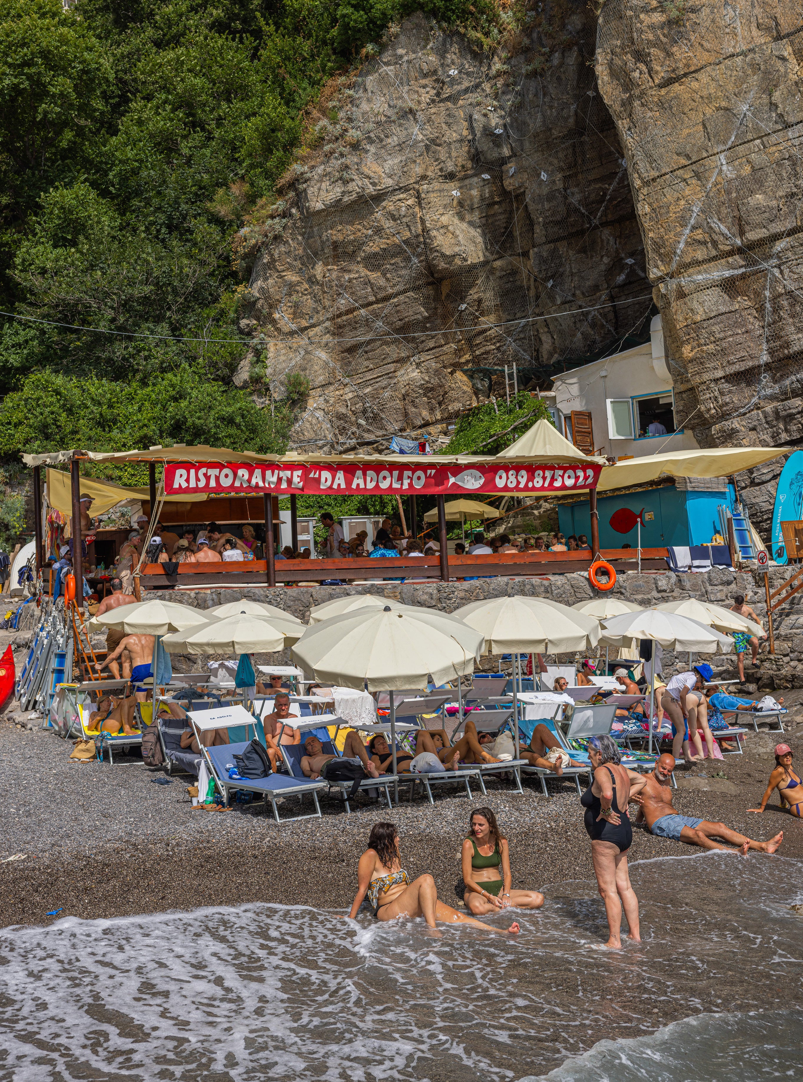 Da Adolfo beach restaurant in Positano with red sign on the building, white sun umbrellas, sunbathers on pebble beach, and cliff face above