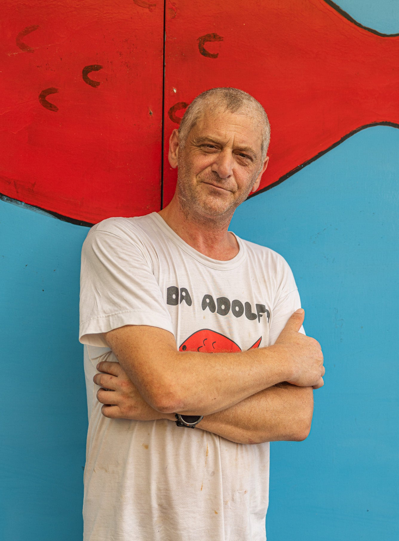 Man in Da Adolfo t-shirt standing with arms crossed in front of a painted red fish mural on a blue wall