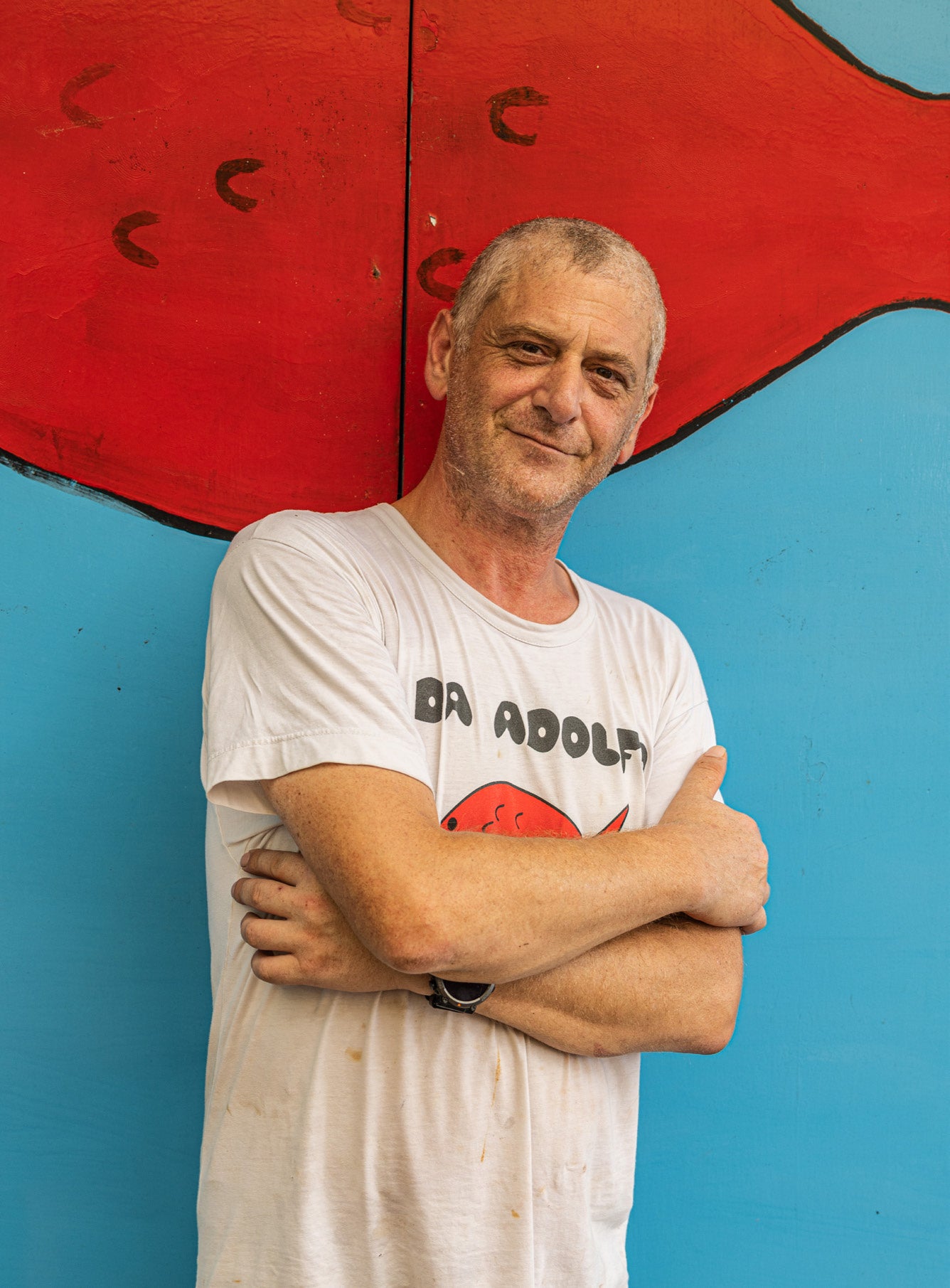 Man in Da Adolfo t-shirt smiling with arms crossed in front of the red fish mural on a blue wall in Positano