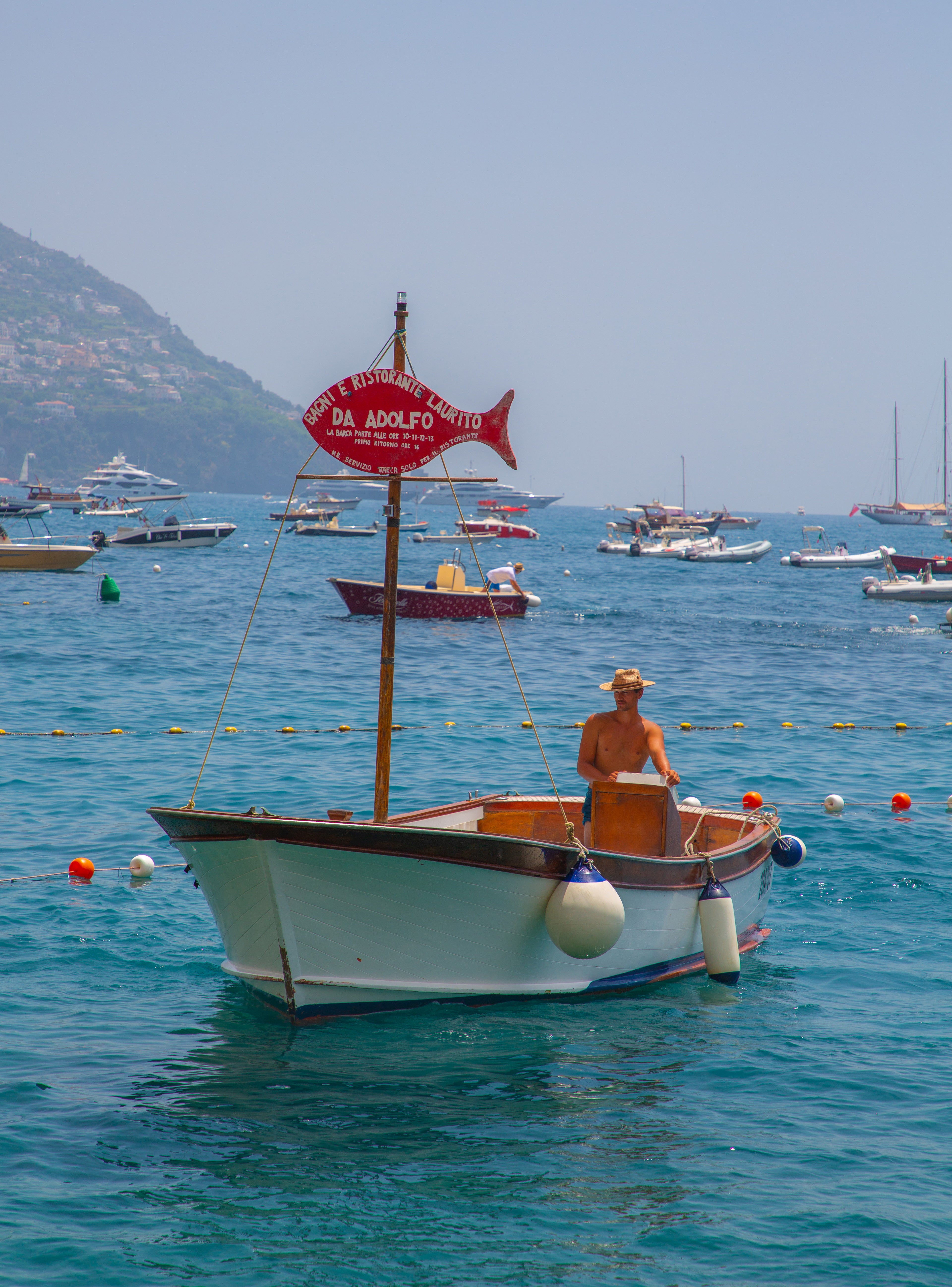 Small white wooden boat with the Da Adolfo red fish sign on a mast in Positano harbour with boats and coastline beyond
