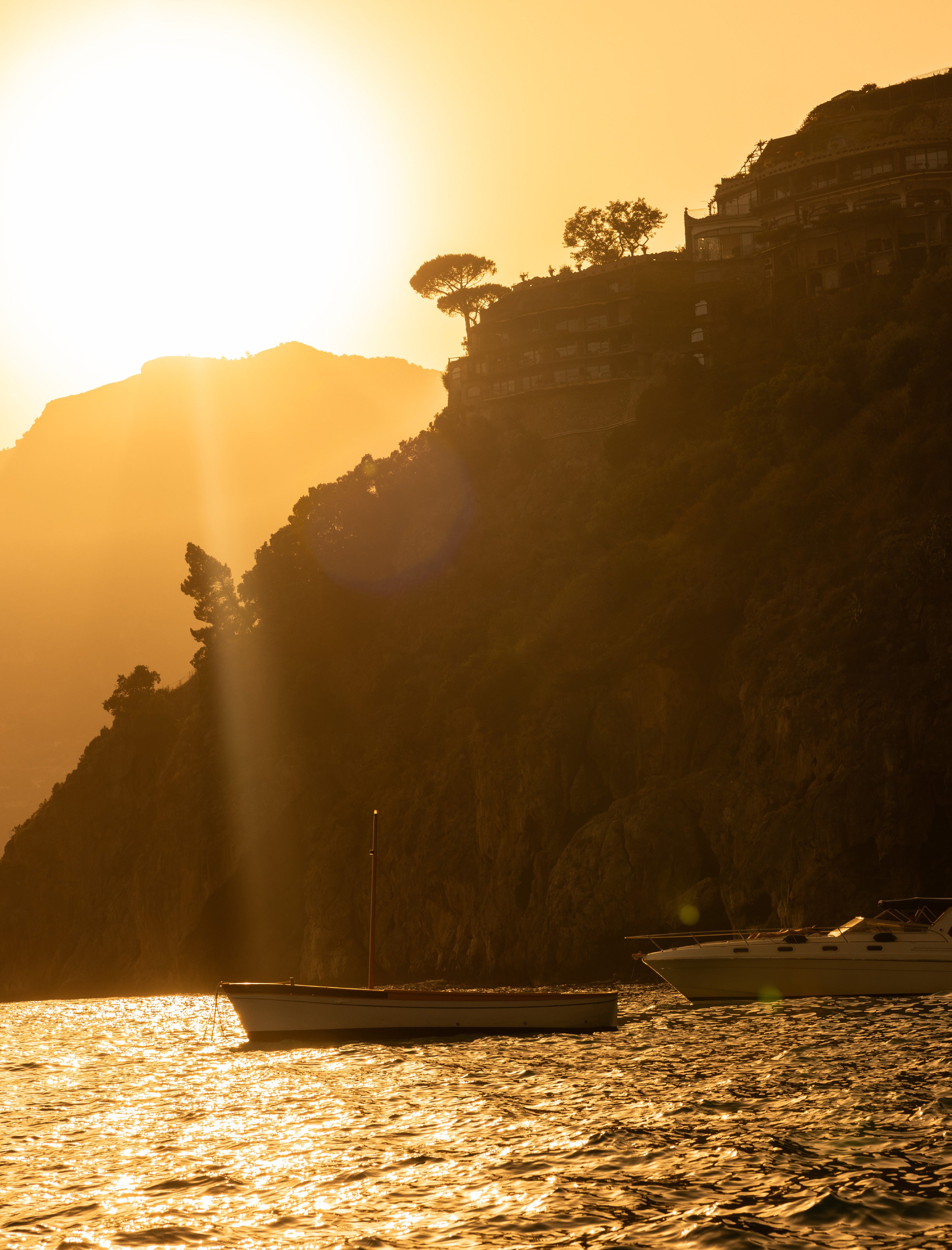 Silhouette of pine trees and cliff-top buildings in Positano against an intense golden sunset with boats on the water below