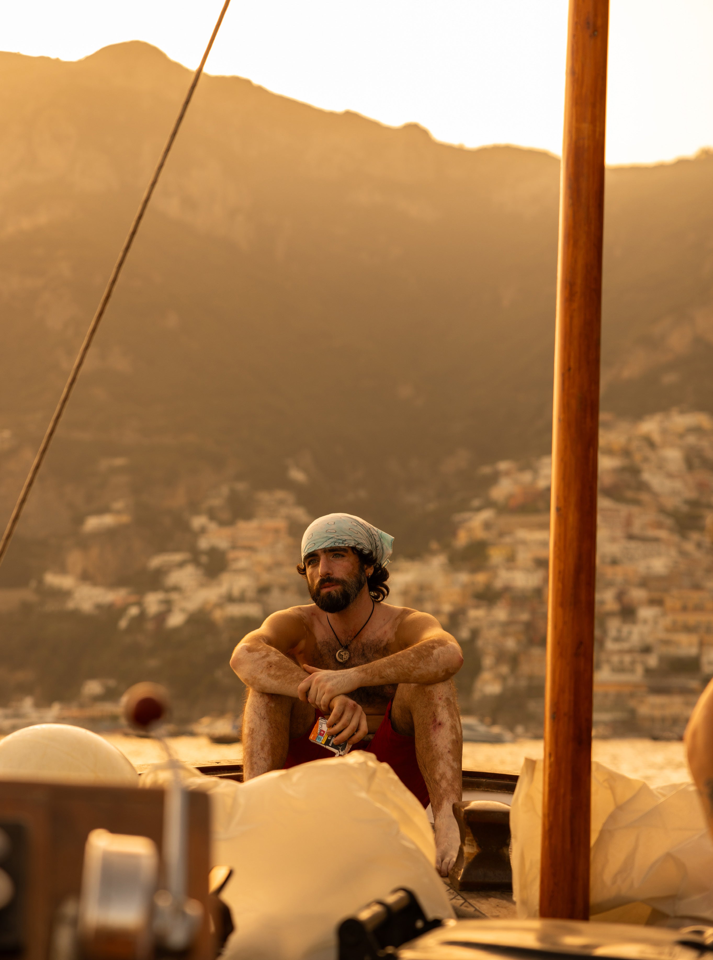 Young man with headband sitting on a boat deck at golden hour with Positano hillside buildings glowing in the background