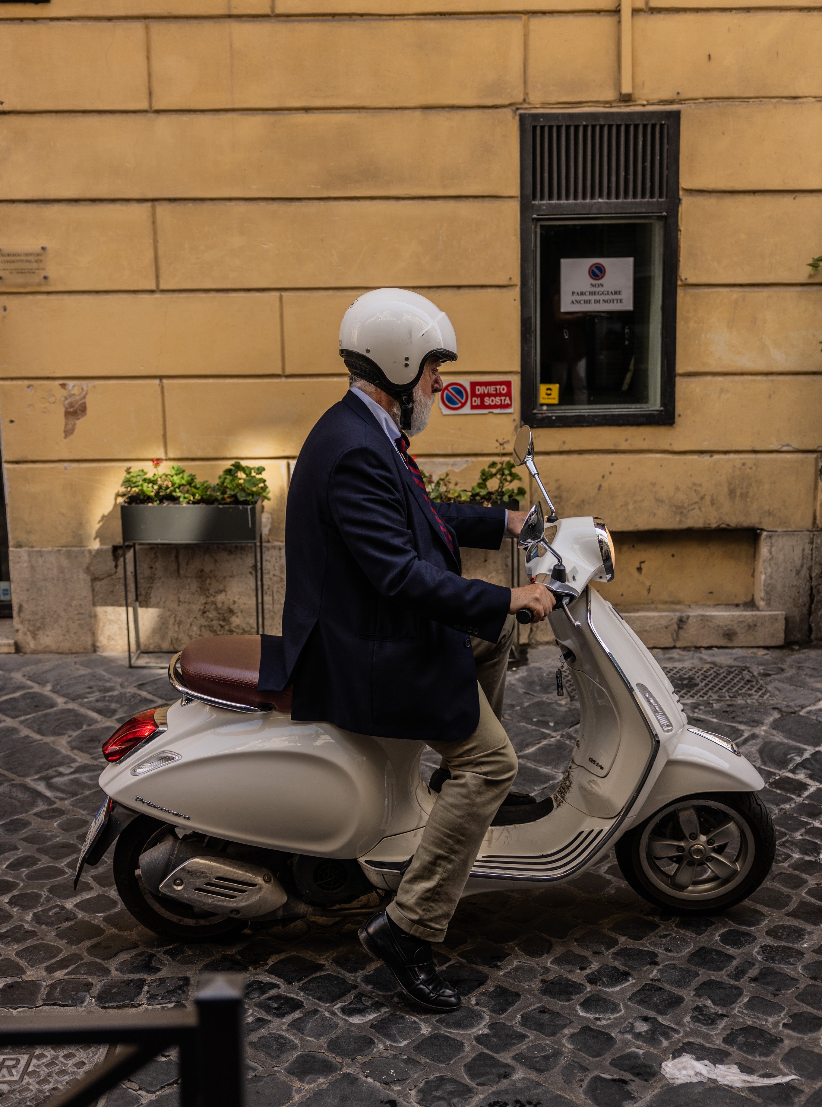 Man in navy suit and white helmet riding a cream Vespa scooter on a cobblestone street in Rome