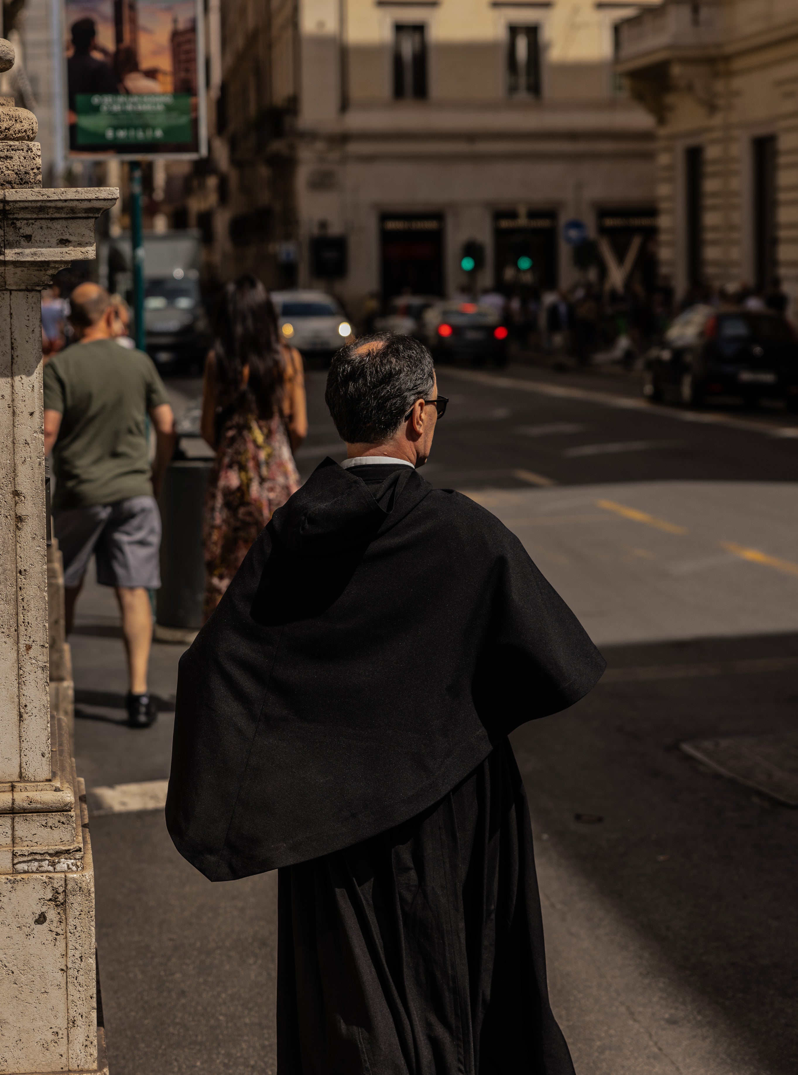 Catholic priest in black cassock walking away from camera on a sunlit street in Rome with pedestrians and traffic in the background