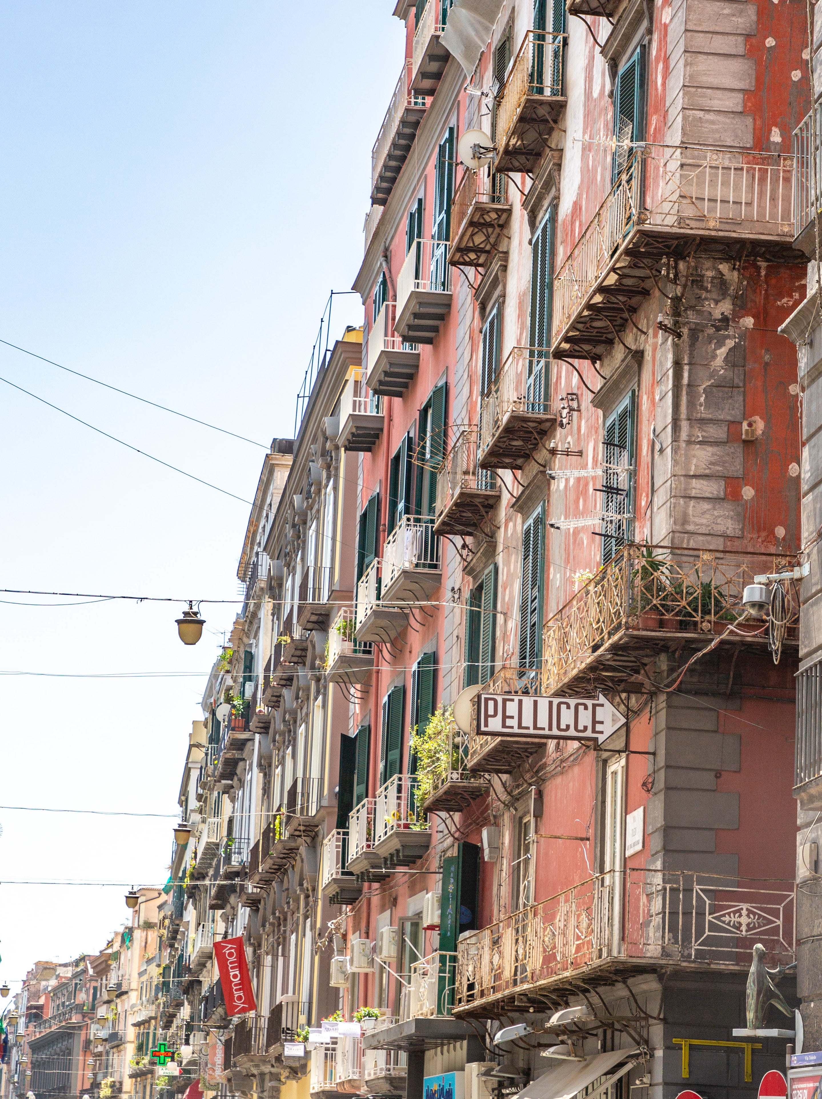 Colourful faded pink apartment buildings with wrought-iron balconies and green shutters lining a narrow street in Naples