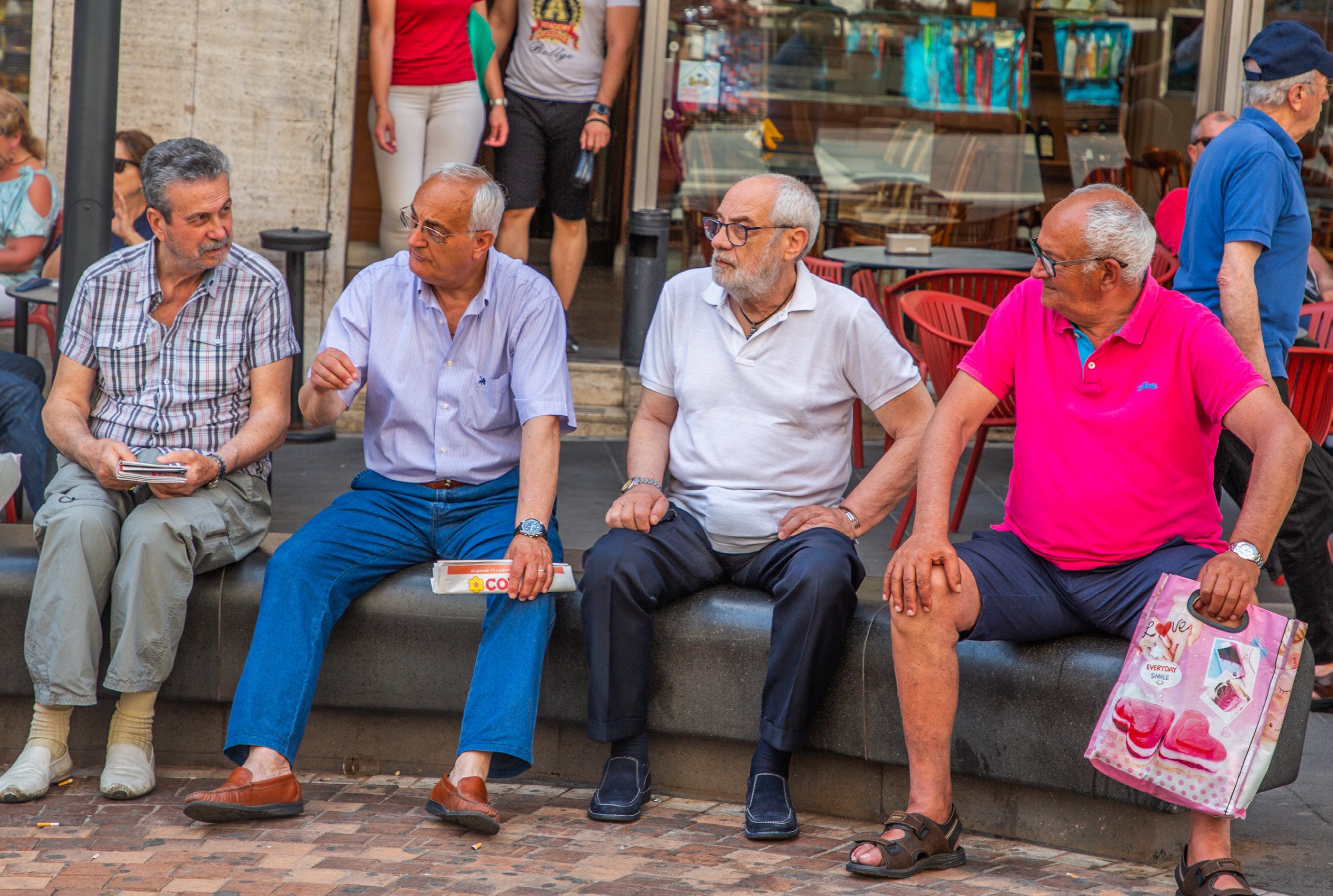 Four older Italian men sitting side by side on a stone bench in a Naples piazza chatting together