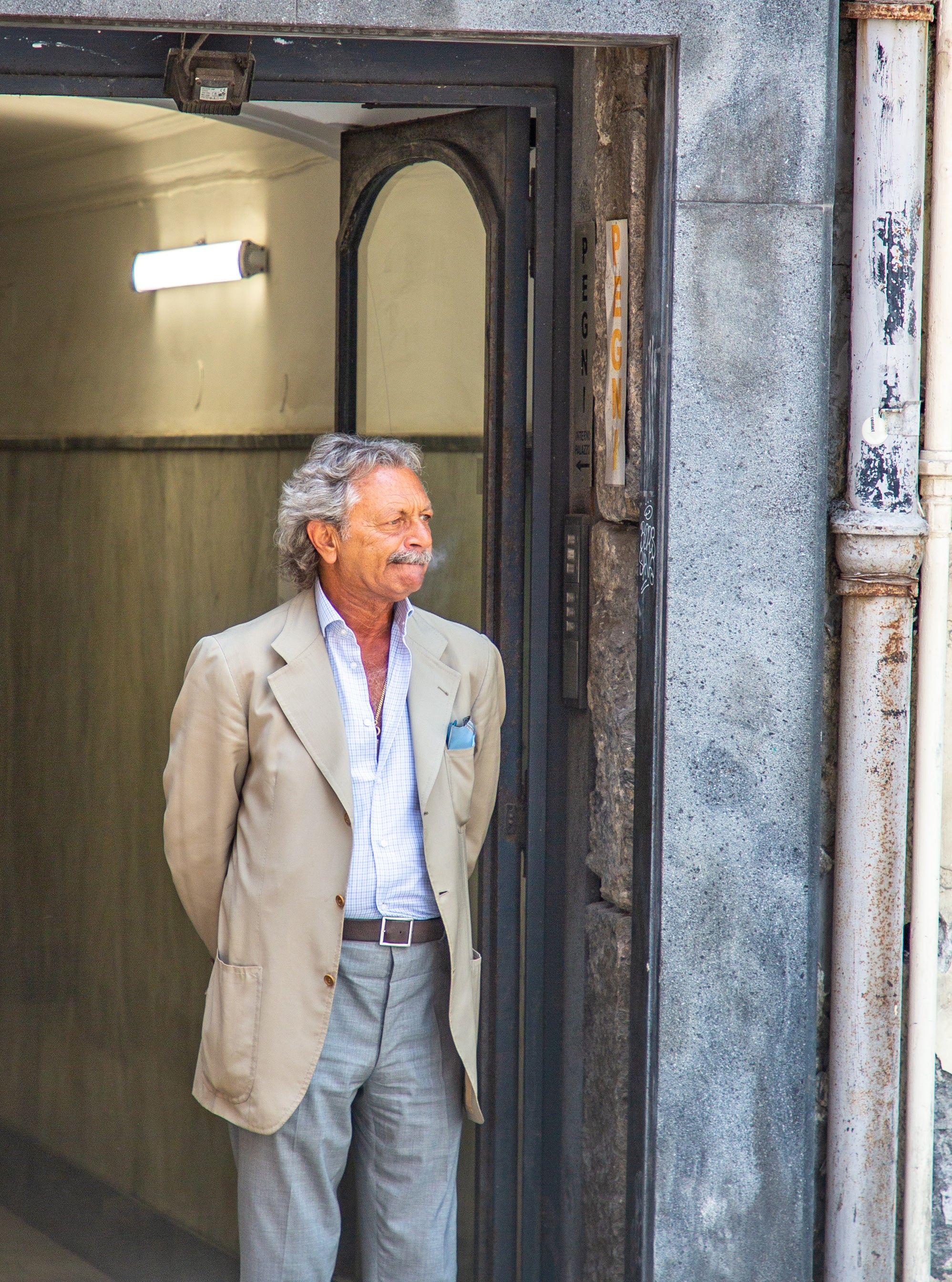 Distinguished older man in a tan blazer and pocket square standing in a worn stone doorway looking out onto a Naples street