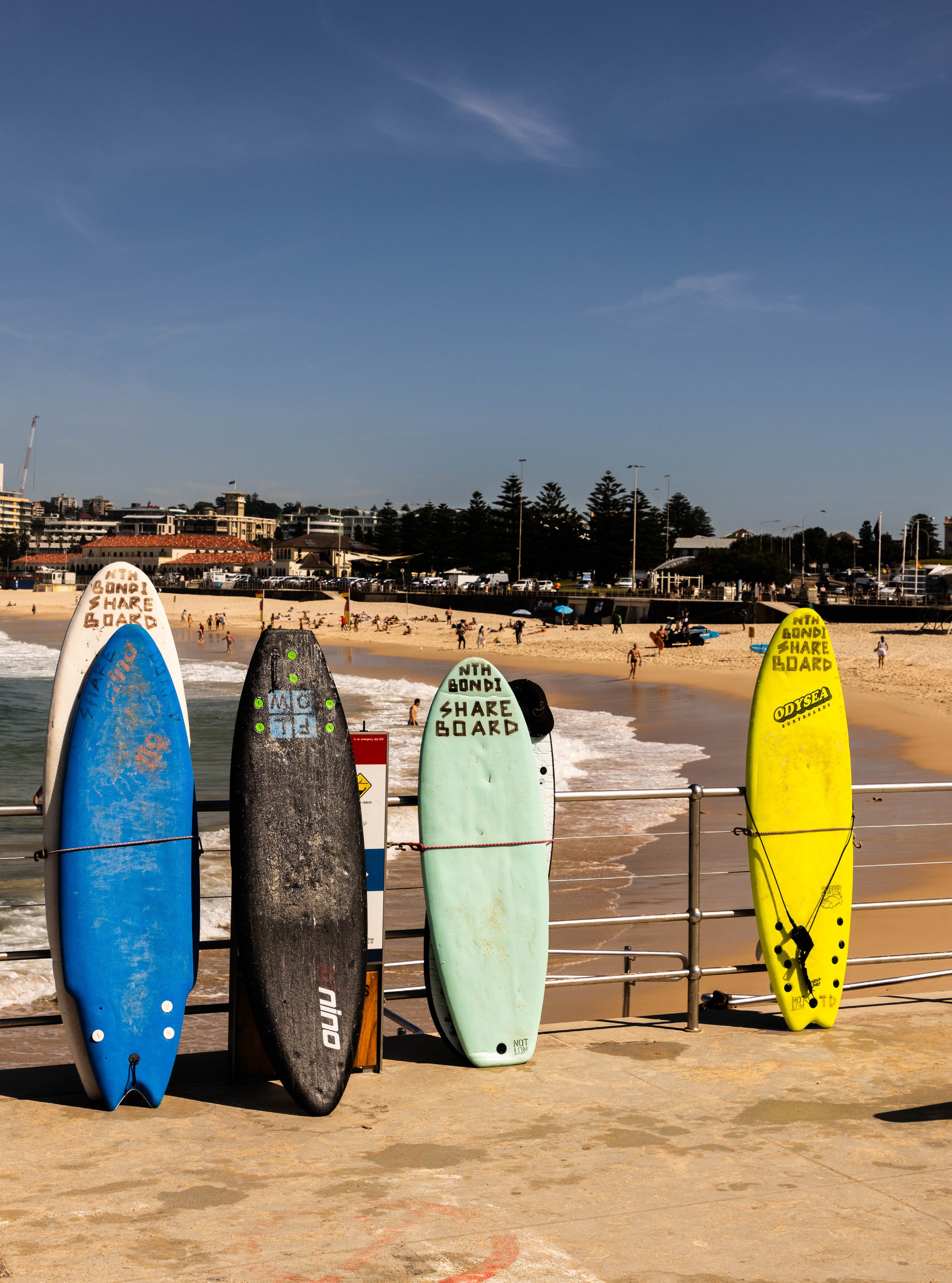 Five colourful surfboards standing upright against a railing at Bondi Beach Sydney with the beach and swimmers behind