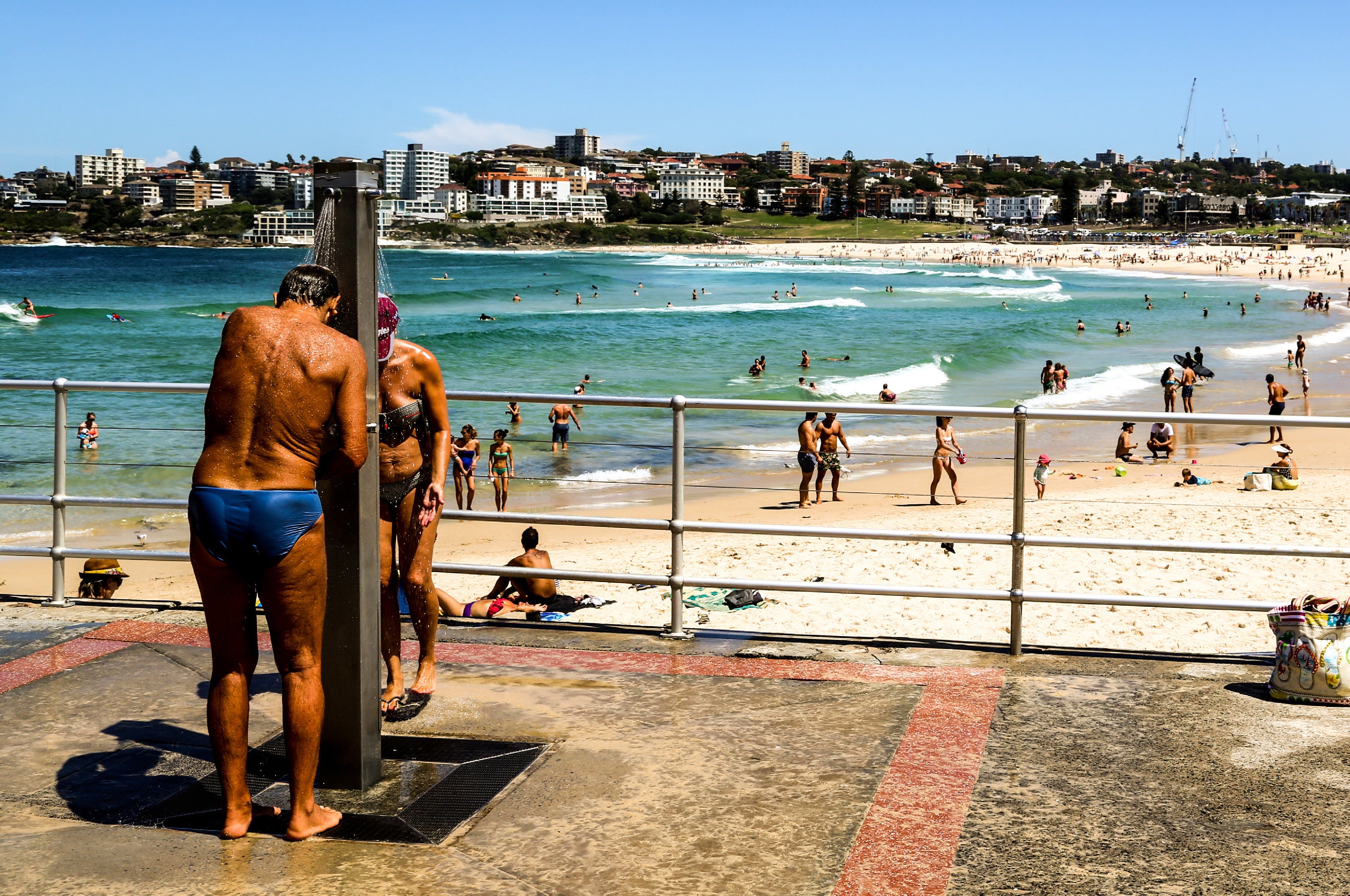 Man rinsing off under an outdoor shower at Bondi Beach promenade with the crowded beach and ocean in the background