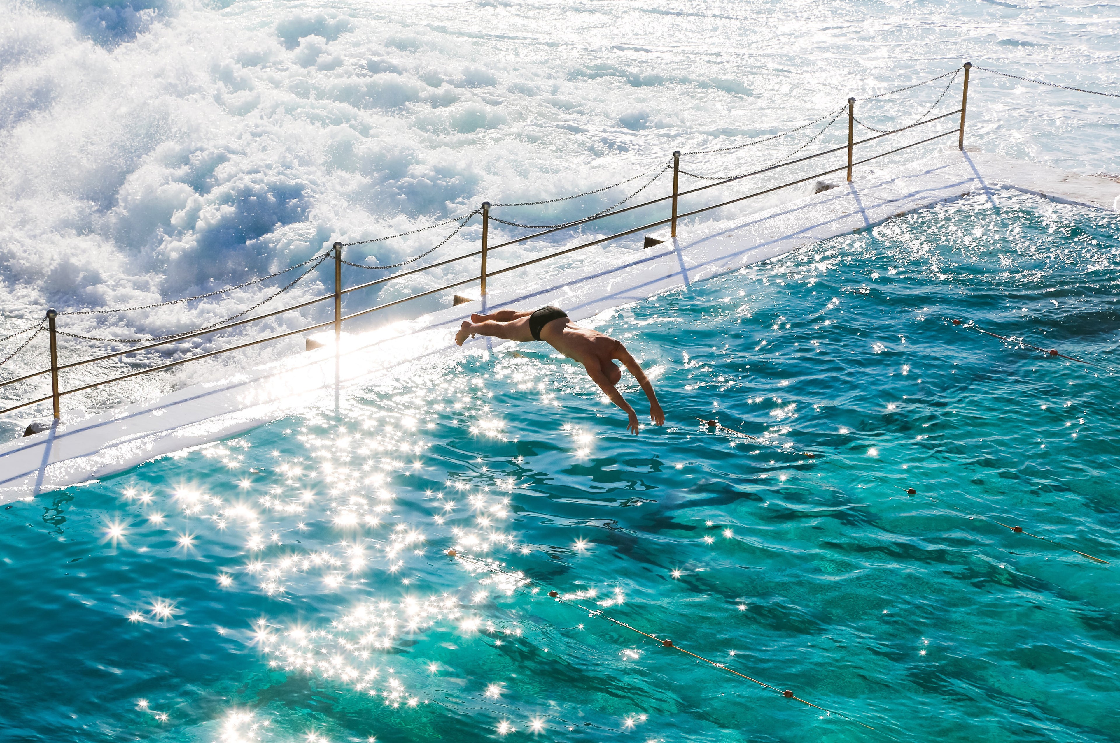 Person diving into sparkling turquoise water at Bondi Icebergs pool with crashing white waves behind the railing