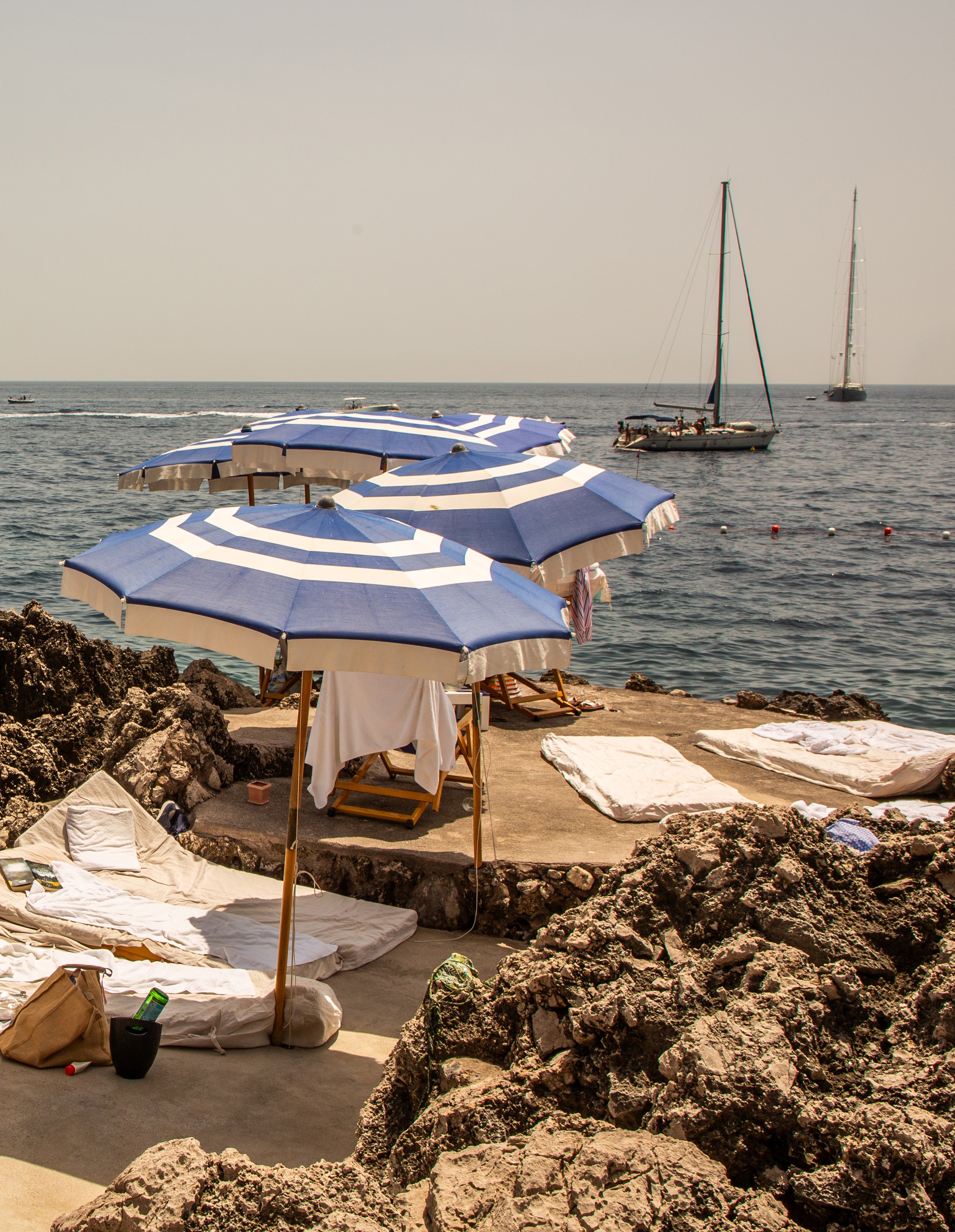 Blue and white striped beach umbrellas on a rocky coastal platform in Capri with sailboats moored in calm waters beyond