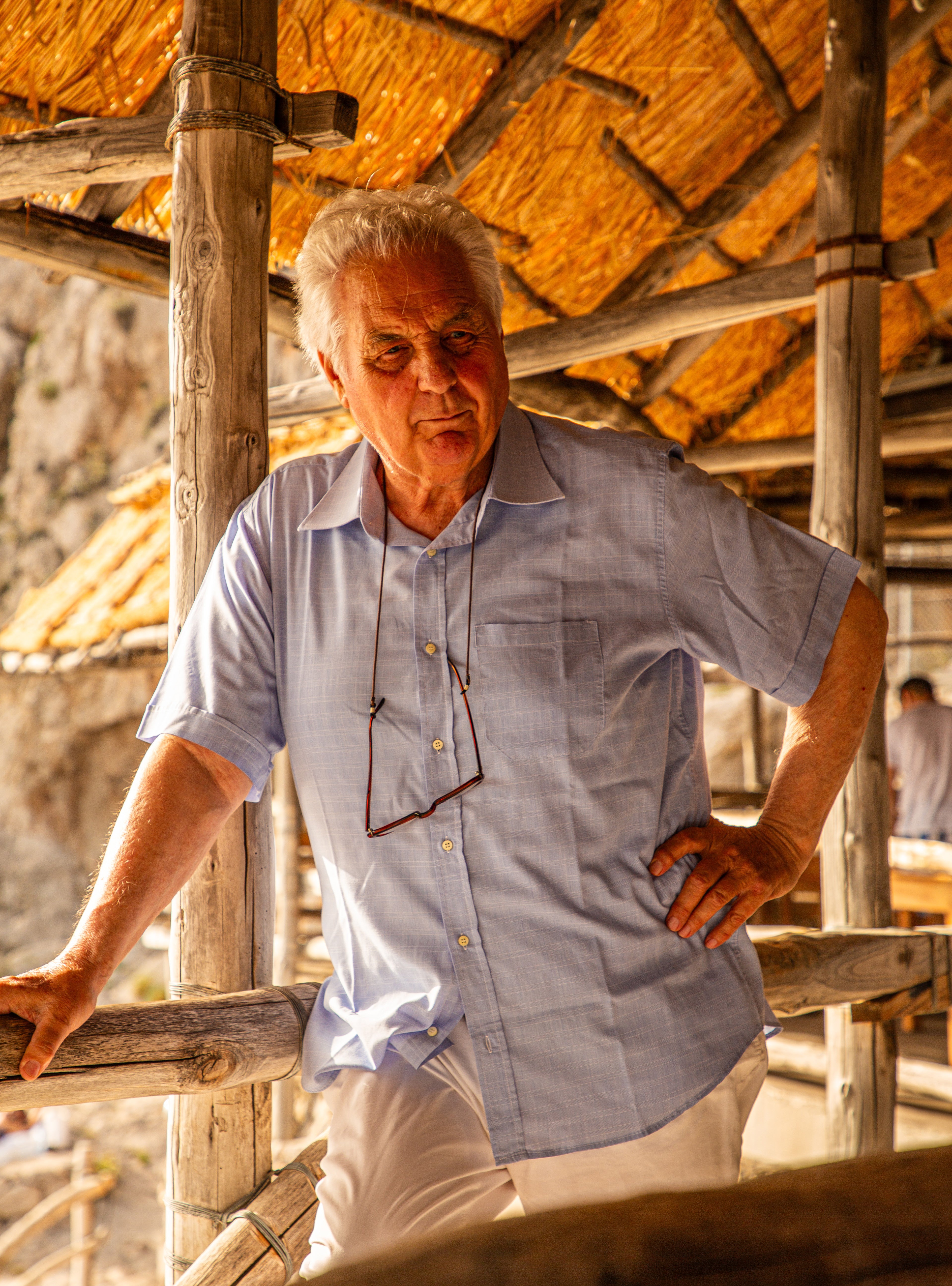 Silver-haired Italian man in a light blue shirt leaning on a bamboo railing beneath a thatched roof shelter in Capri