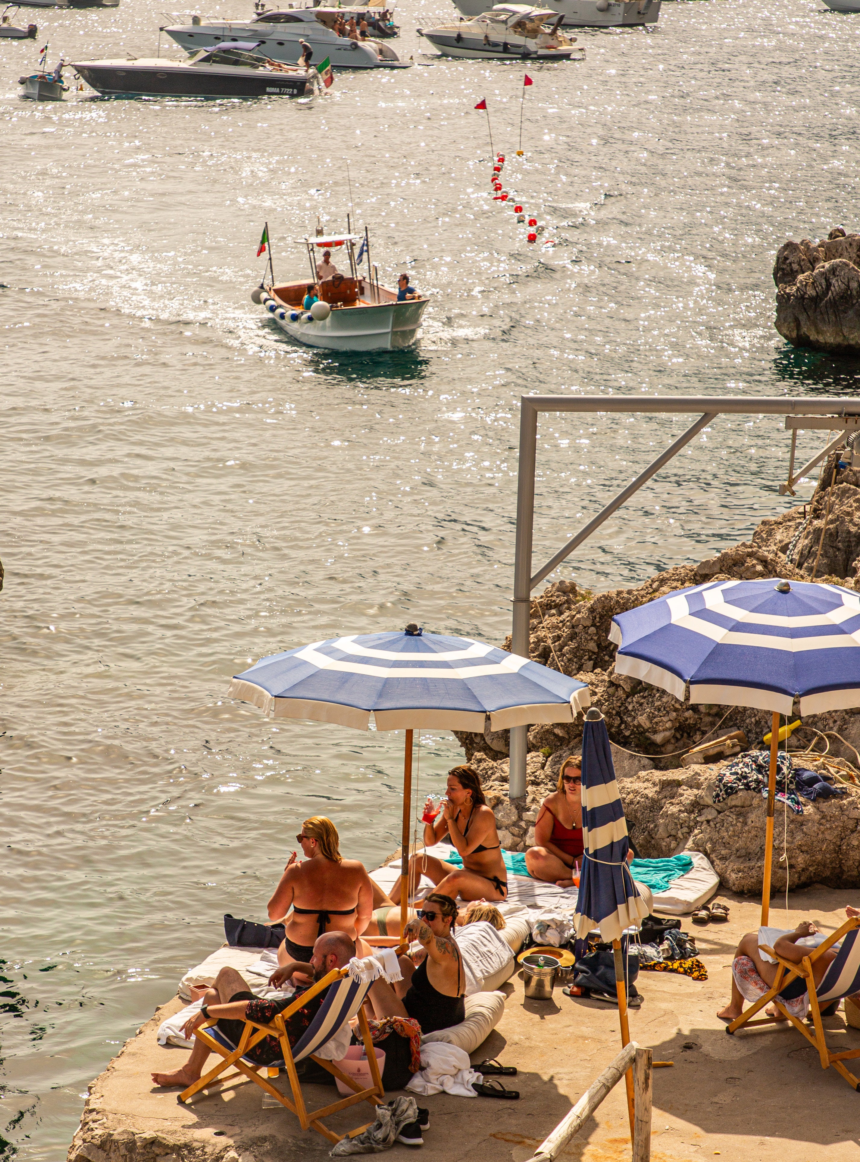Overhead view of sunbathers on a rocky coastal platform under blue and white umbrellas with sparkling water and boats in the harbour beyond