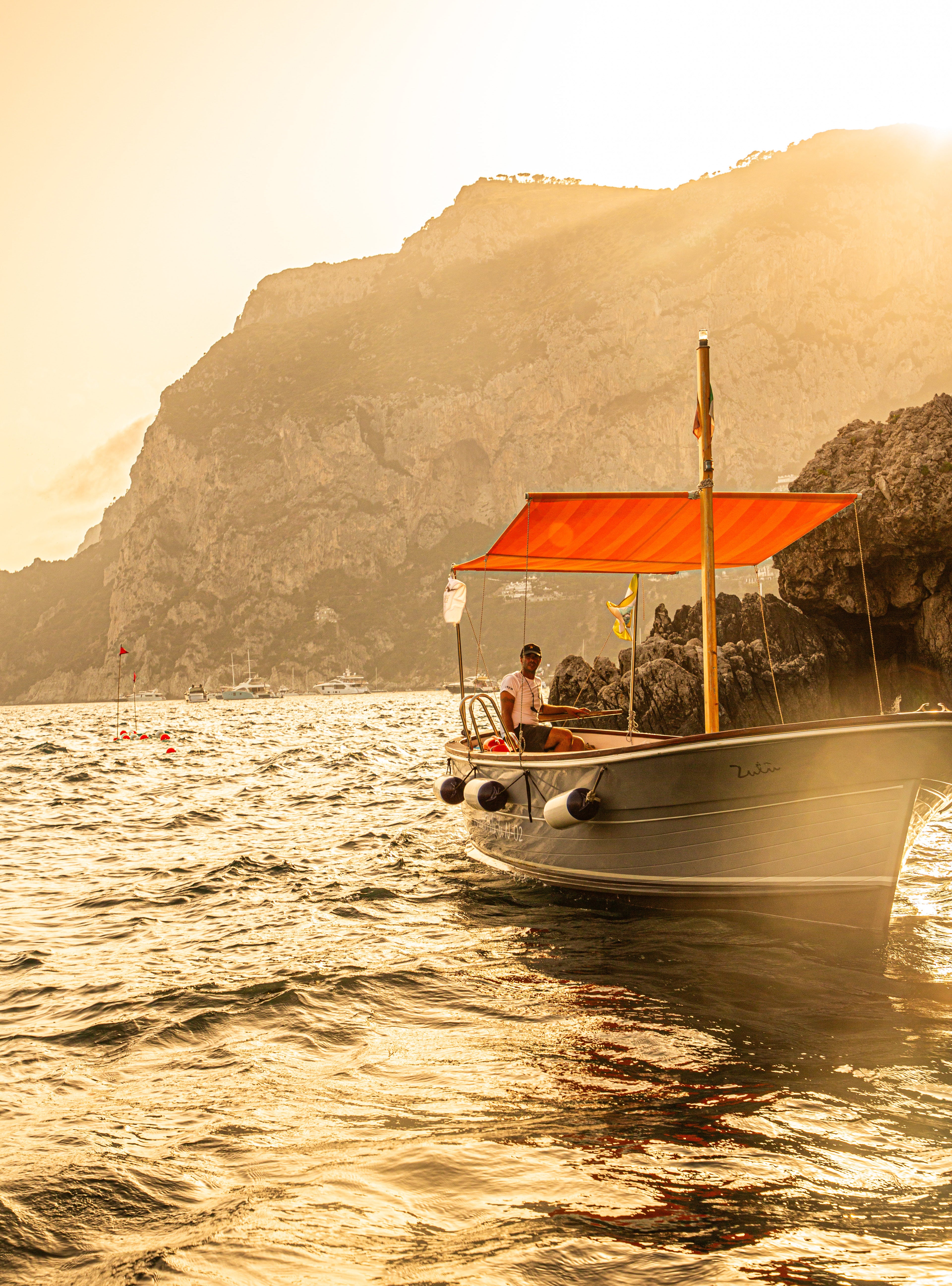 Small boat with orange canopy on golden sunlit water with towering Capri cliffs backlit by the setting sun