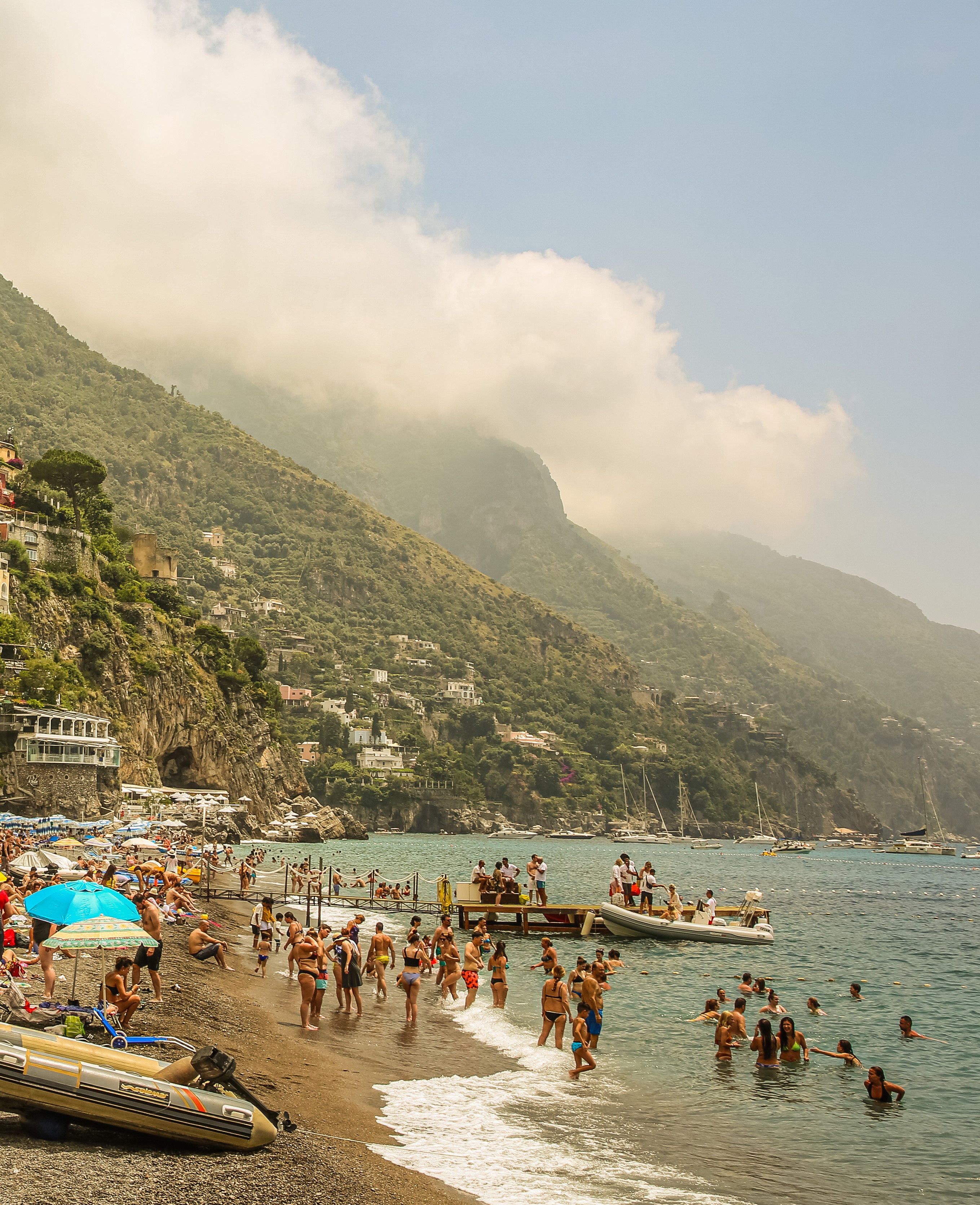 People swimming and wading on a sandy beach in Positano with lush green mountains and low clouds along the Amalfi coastline