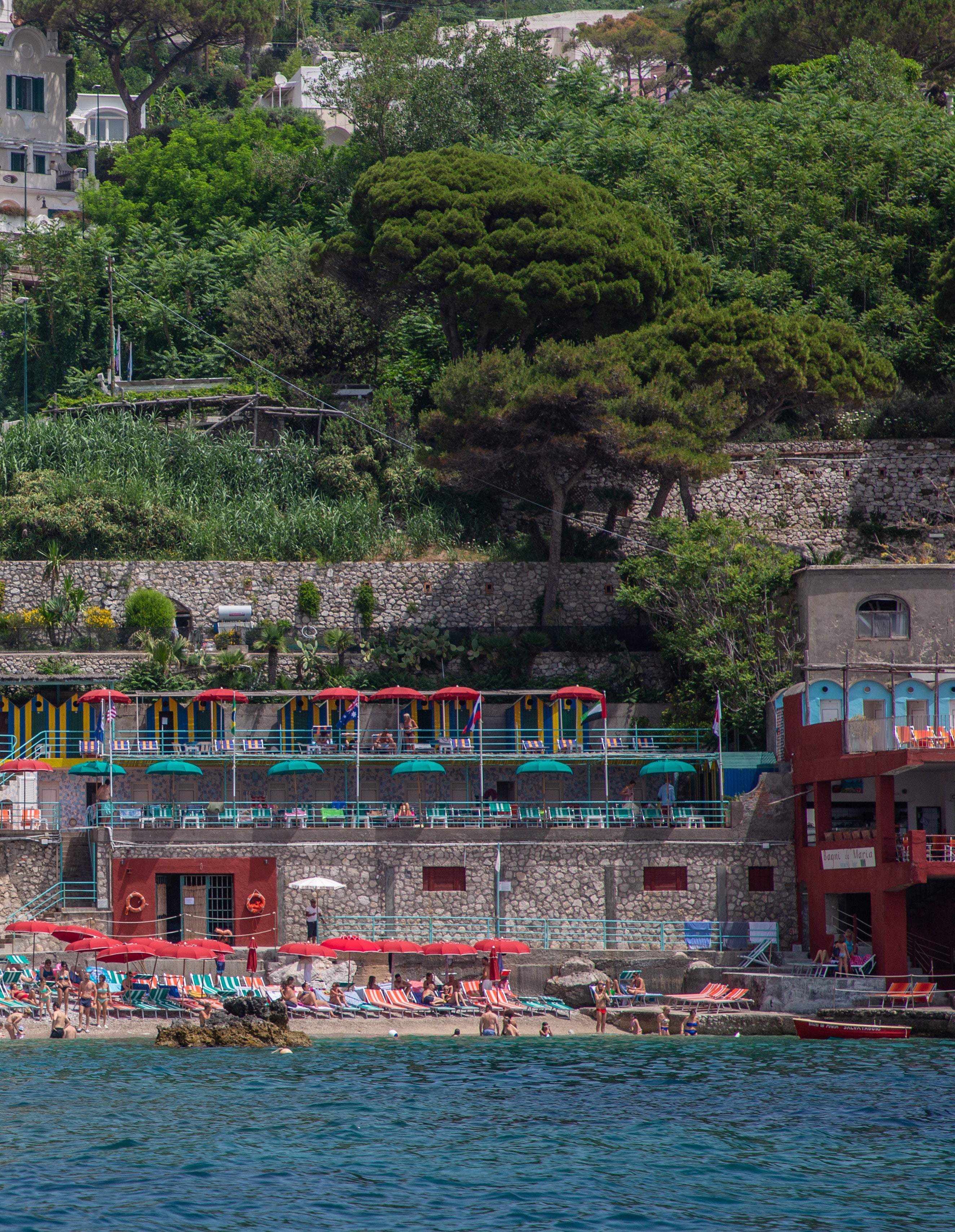 Small beach club with coloured umbrellas built into the base of a lush green cliff on the Amalfi Coast with swimmers below