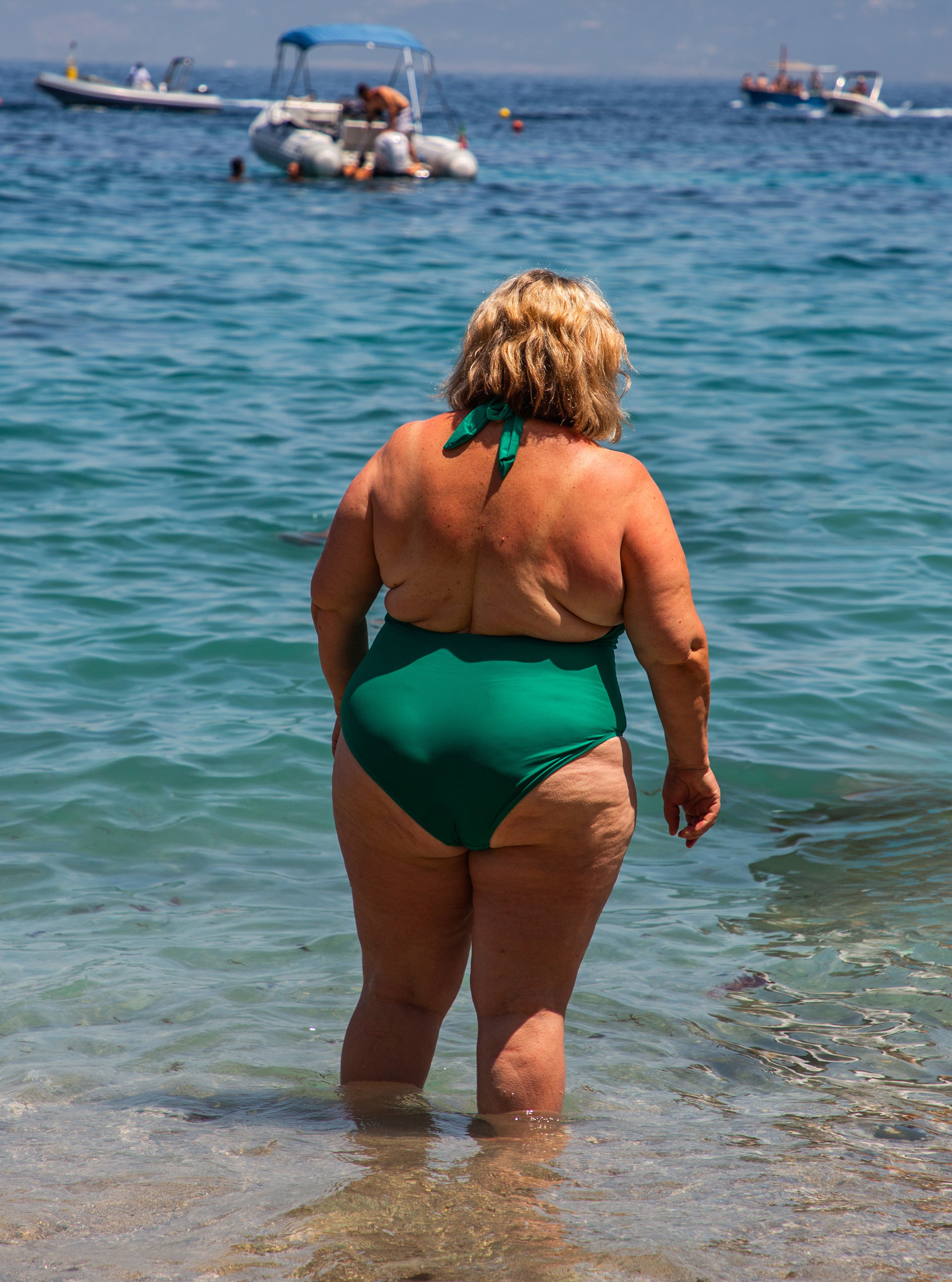 Woman in a green swimsuit seen from behind wading into clear turquoise Mediterranean water with boats anchored in the distance