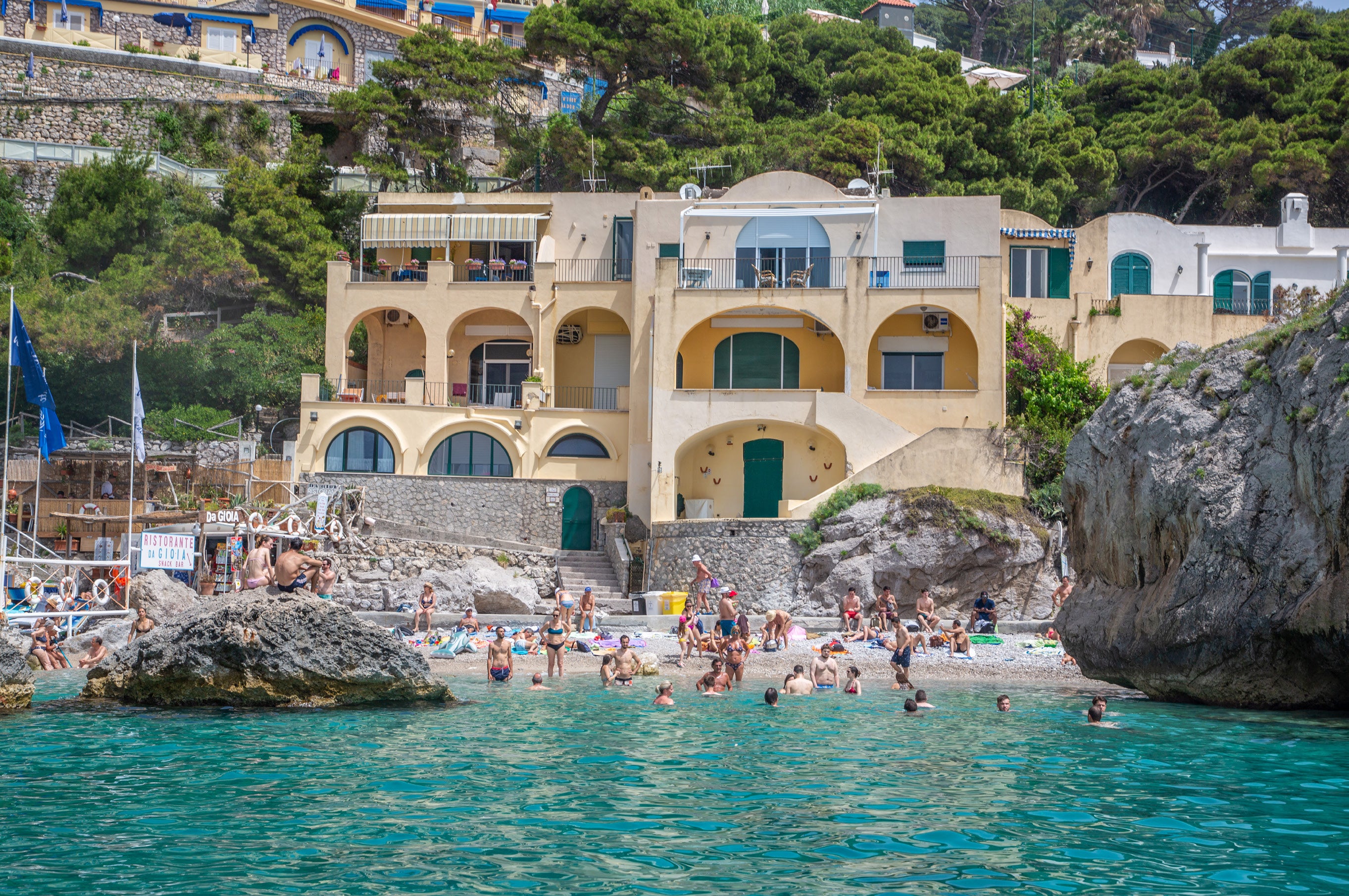 Swimmers in a turquoise cove at the base of yellow Mediterranean villas with arched facades built into rocky cliffs on Capri