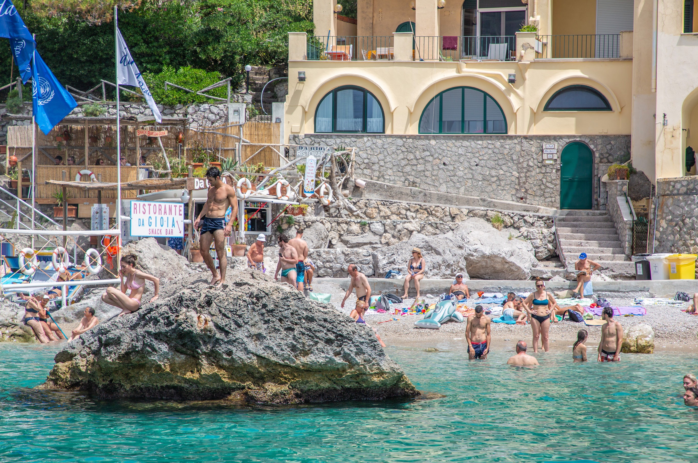 Young man in swim trunks standing on a large rock above turquoise water with swimmers and a beachside restaurant sign in Capri