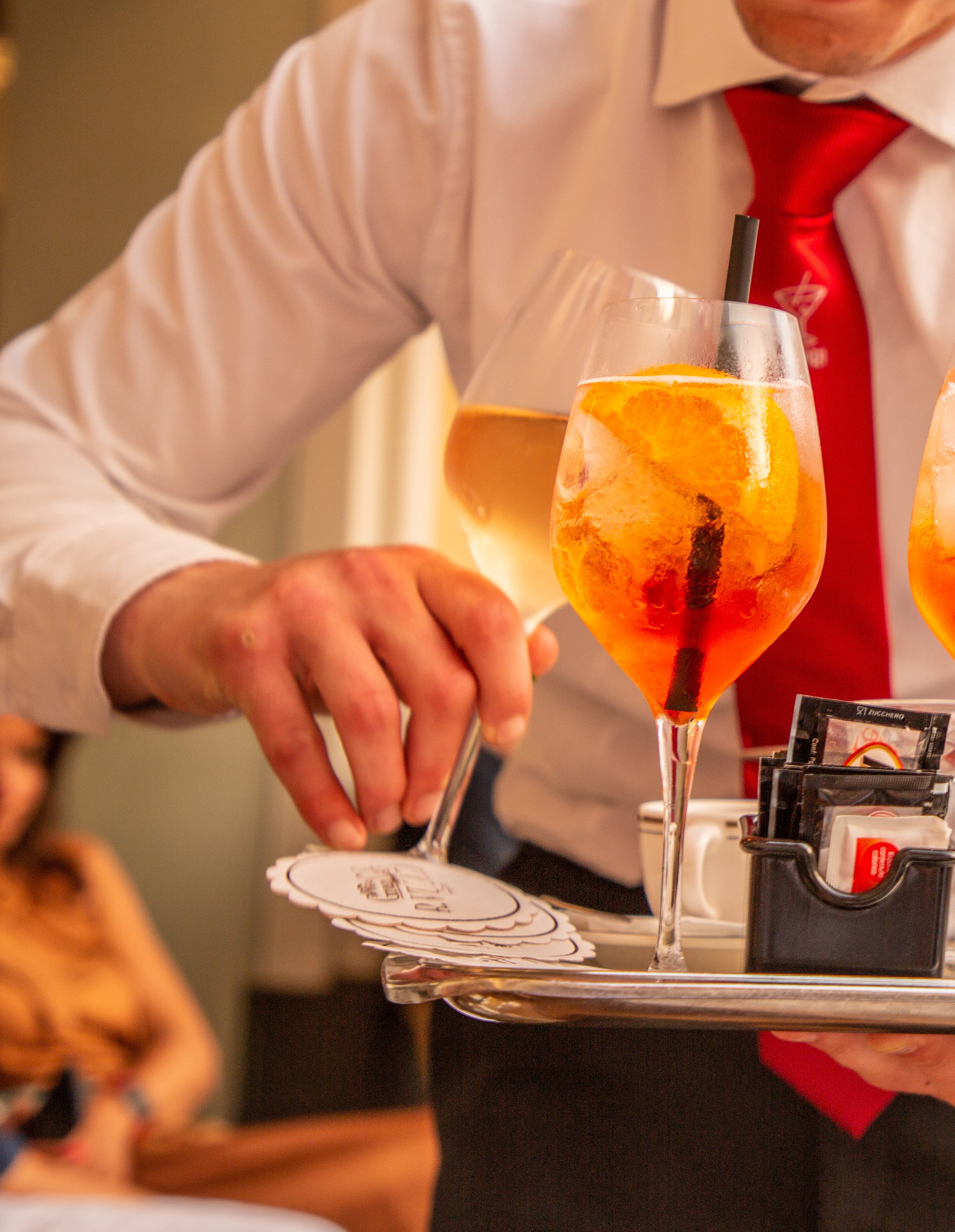 Close-up of a waiter in a white shirt and red tie carrying a silver tray with two golden Aperol spritz cocktails in wine glasses