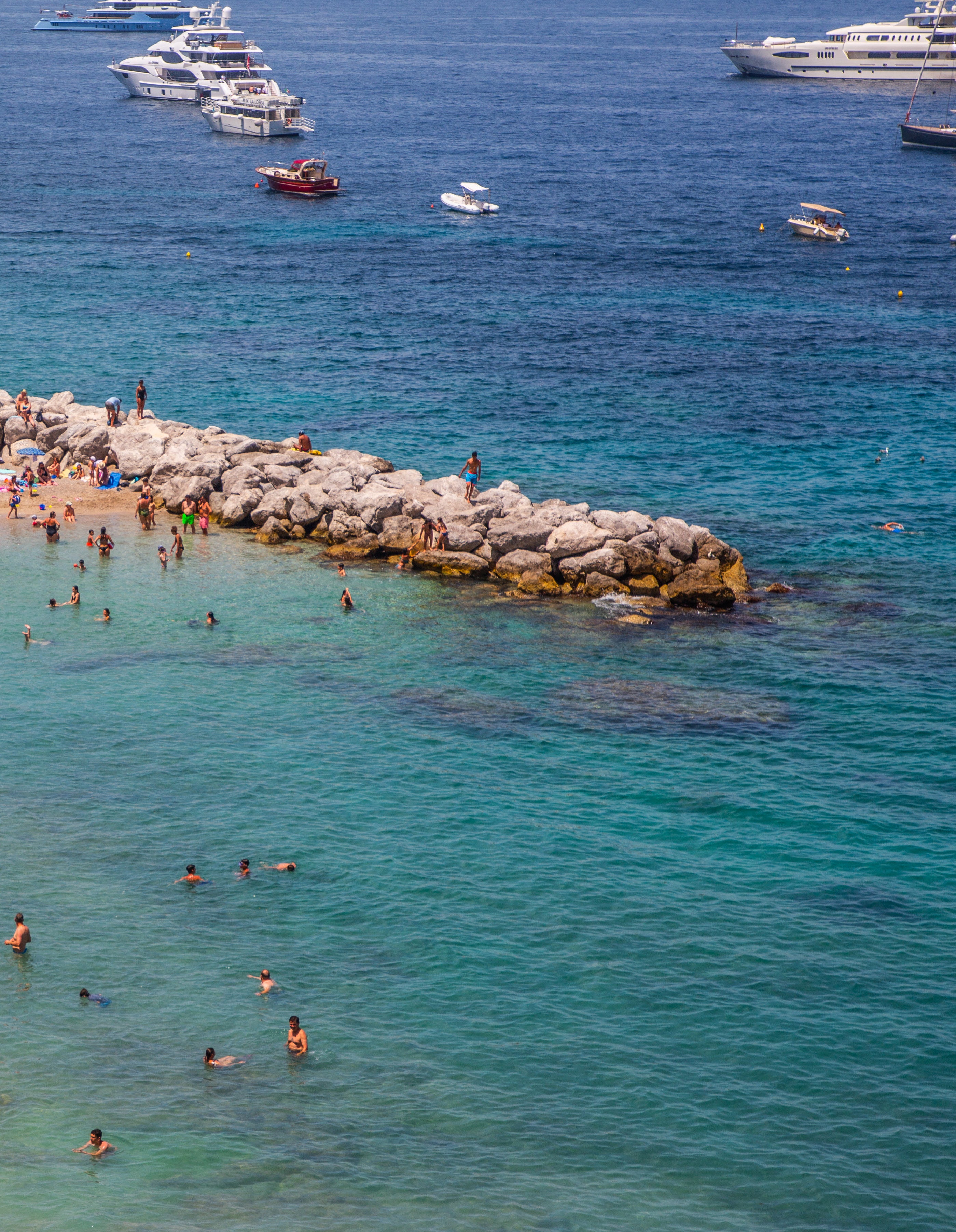 Aerial view of swimmers in brilliant turquoise water around a rocky breakwater jetty with white yachts anchored in the deep blue sea beyond