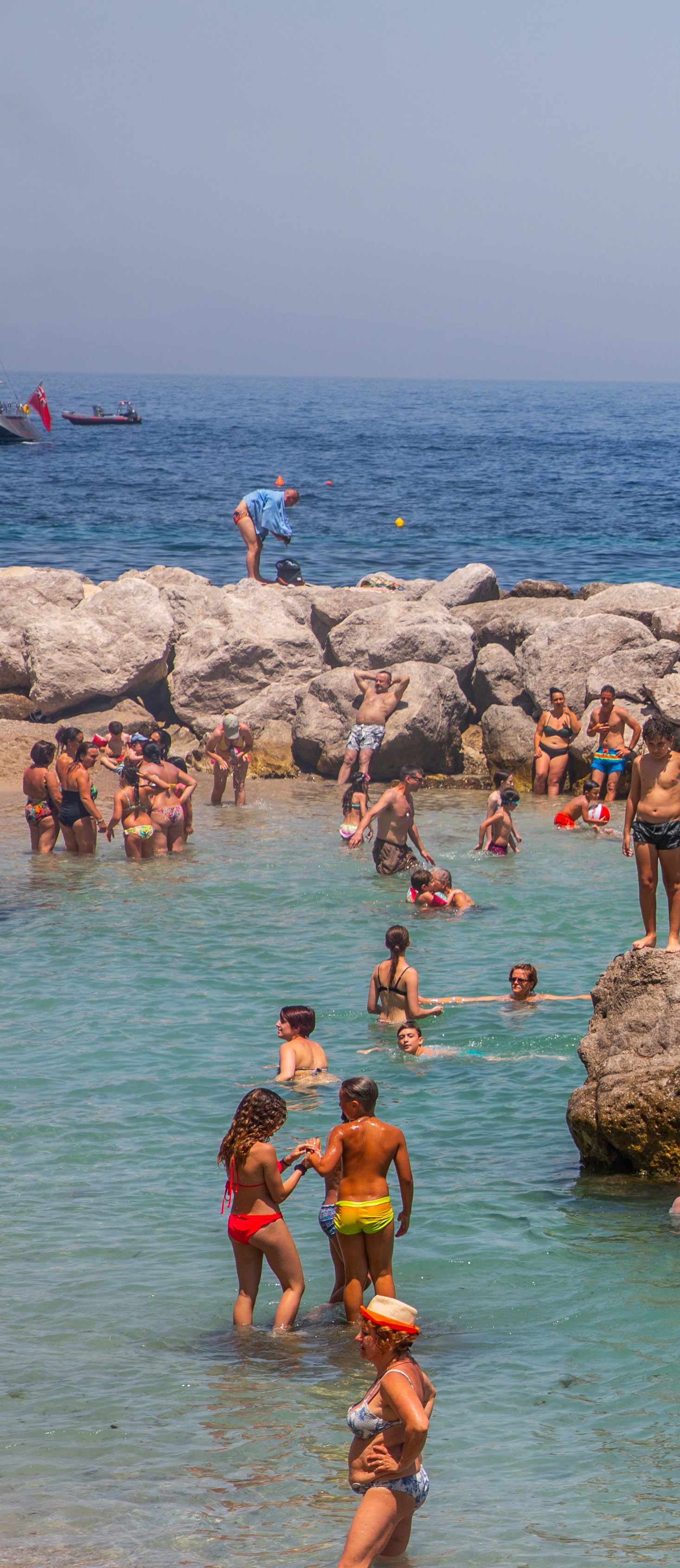 Beachgoers wading and swimming in shallow turquoise water among large sun-bleached rocks on the Italian coast on a hazy summer day