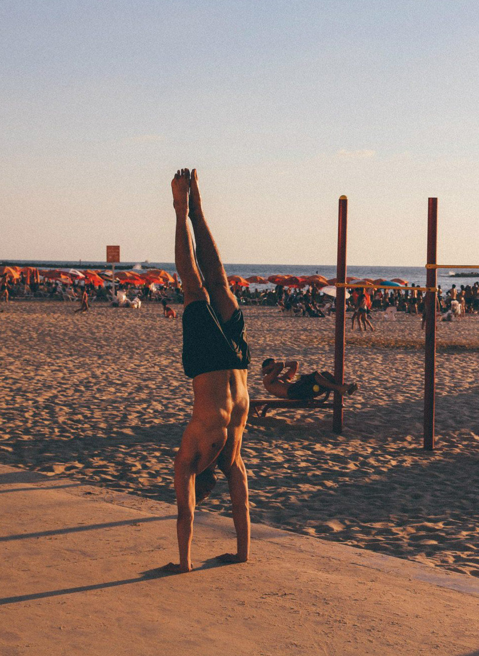 Man performing a perfect handstand on sandy beach beside outdoor fitness bars at golden hour with beachgoers and sea in the background