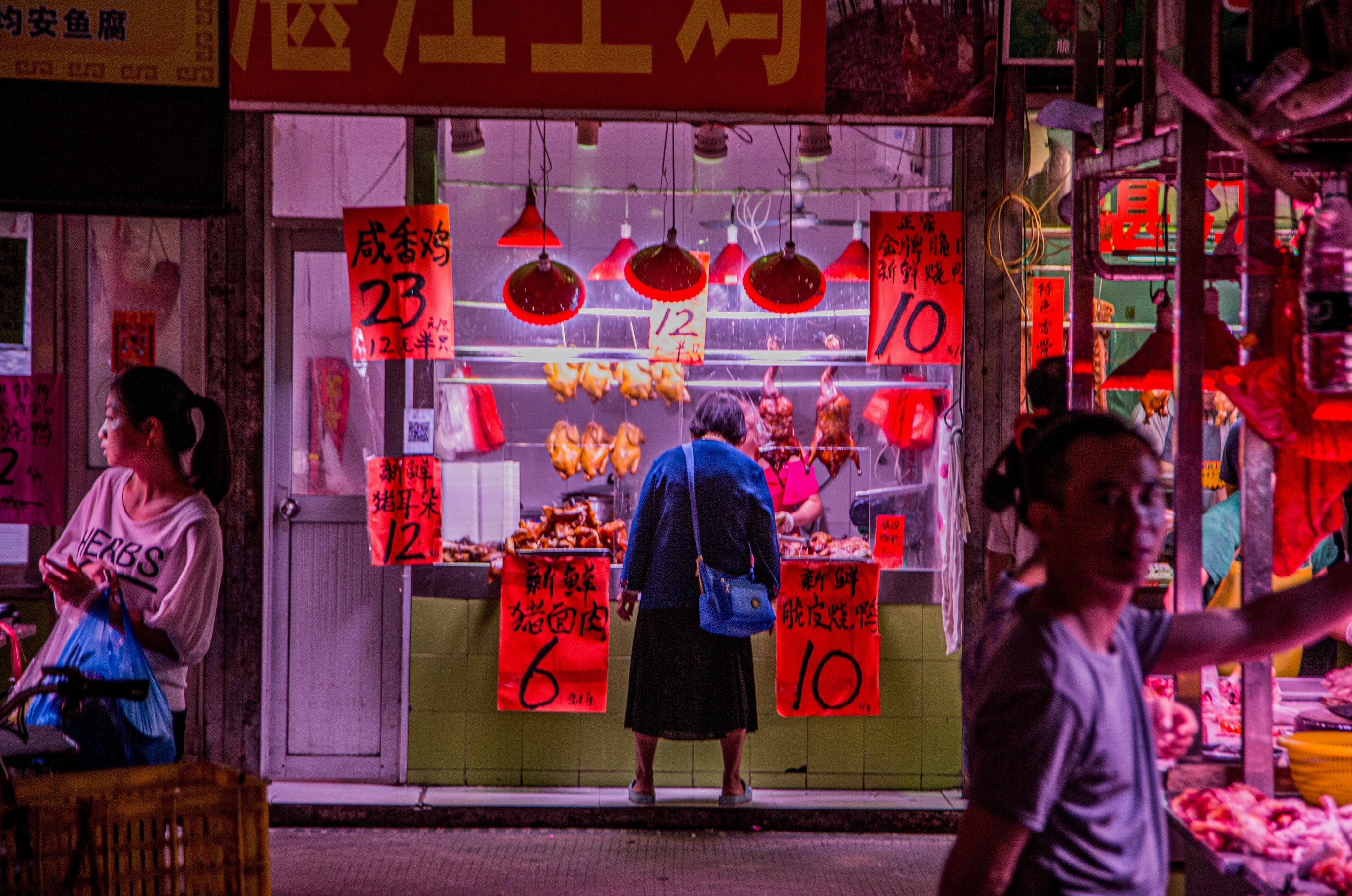 Customer browsing a warmly lit butcher shop at night with red pendant lamps illuminating hanging roast poultry and hand-painted price signs