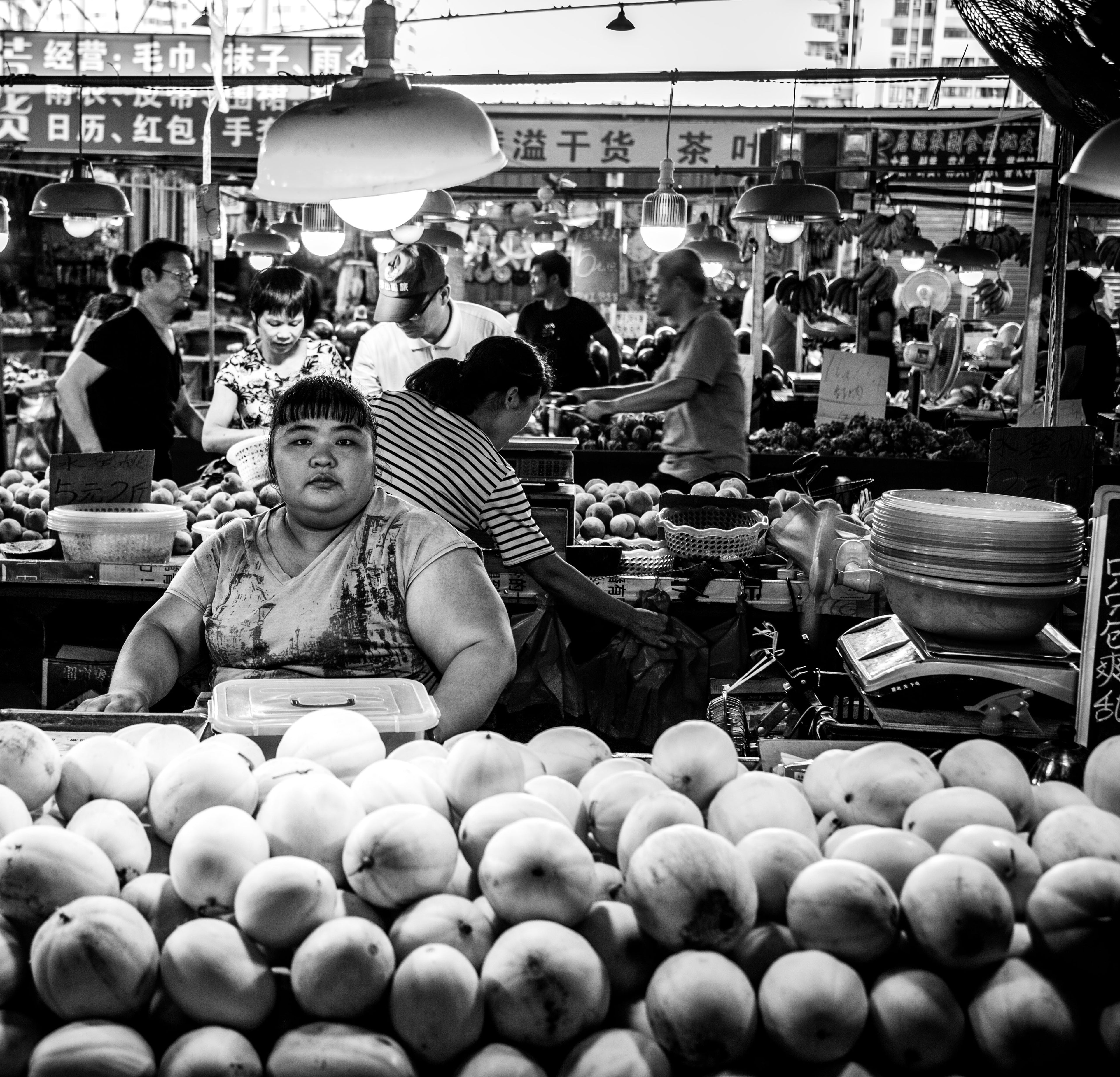 Black and white portrait of a woman sitting behind a large display of fresh round peaches at a crowded open-air market with overhead lights